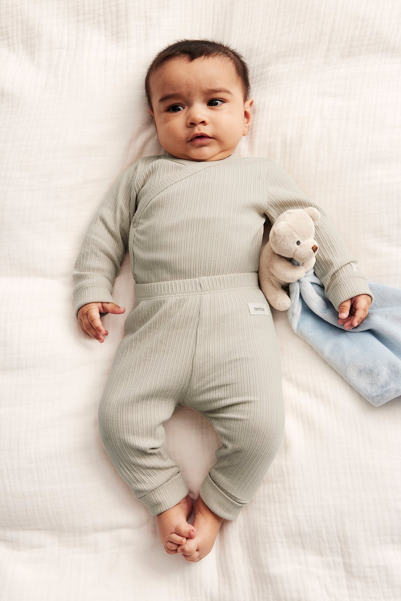 Top-down view of a baby wearing beige ribbed leggings with a matching wrap top, holding a teddy bear comforter.