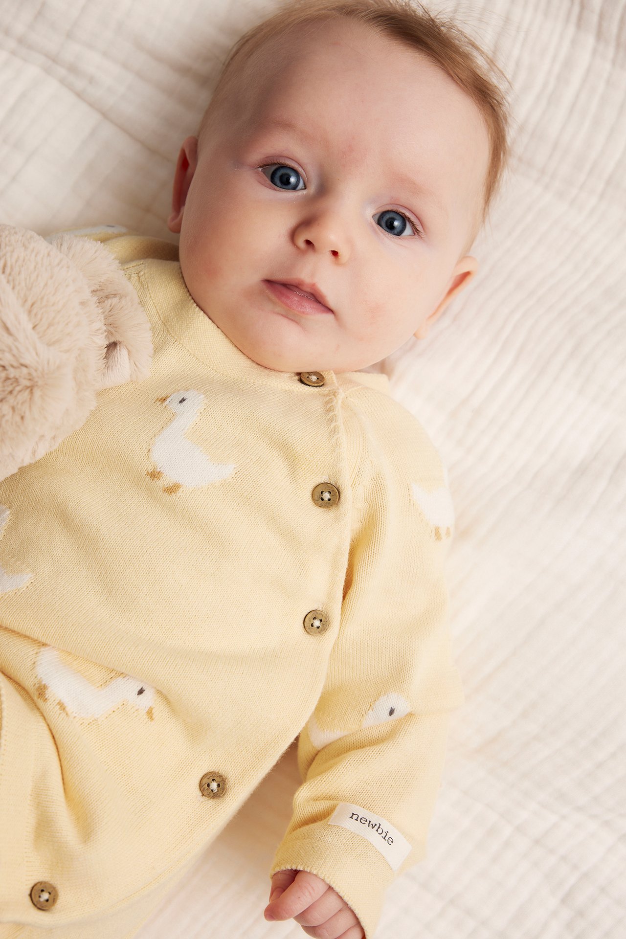 Close-up of a baby in a yellow knitted cotton cardigan with duck embroidery, side buttons, holding a plush toy.