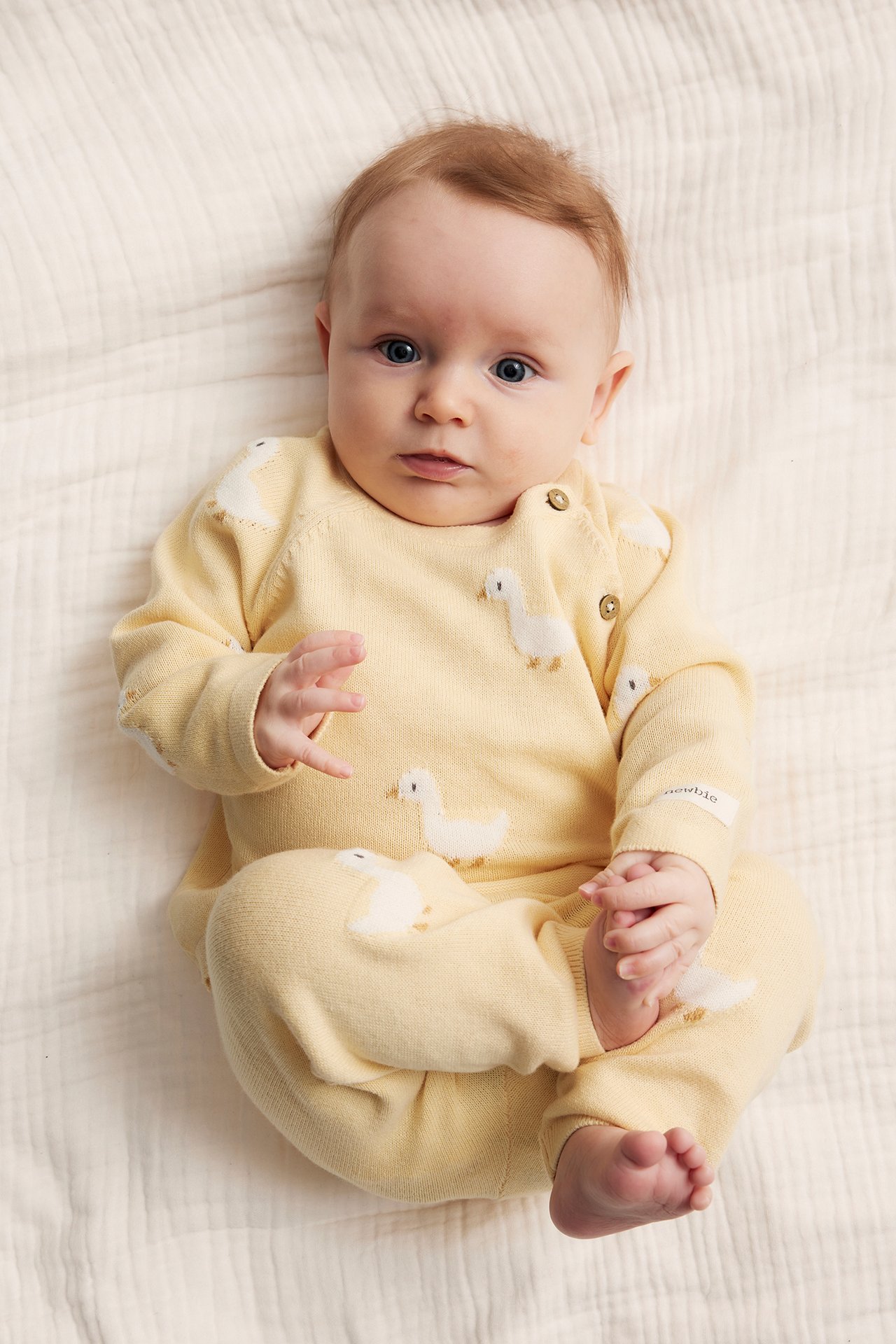 Top-down view of a baby in a yellow knitted cotton set with white duck embroidery, side buttons.