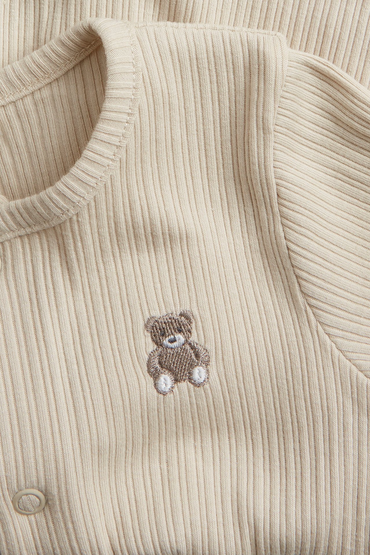 Close-up front view of a beige ribbed baby pyjama with snap buttons and a teddy bear embroidery.