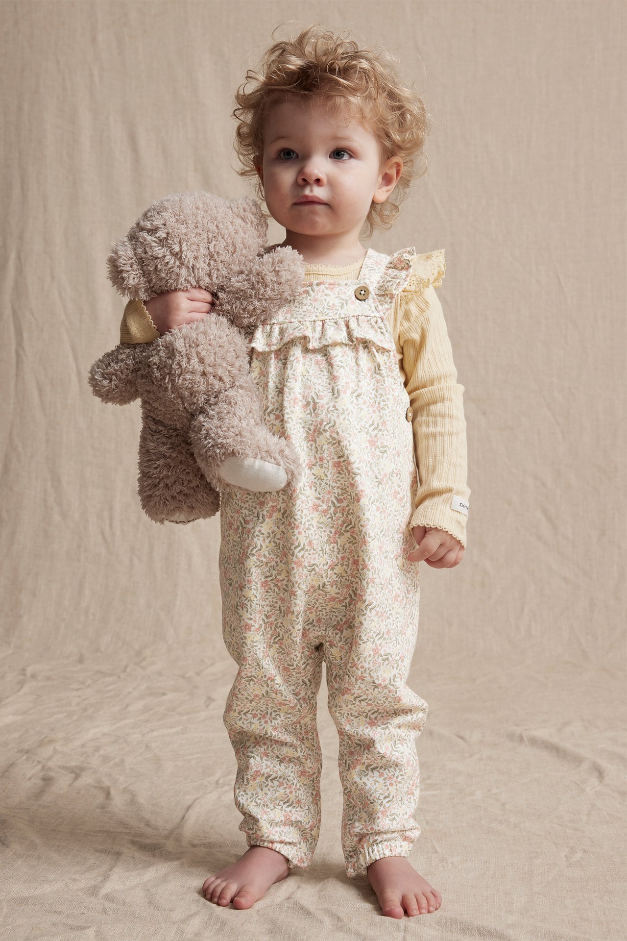 Vorderansicht: Baby in cremeweißer geblümter Baumwoll-Latzhose mit Rüschen über gelbem Oberteil, hält einen braunen Teddy.
