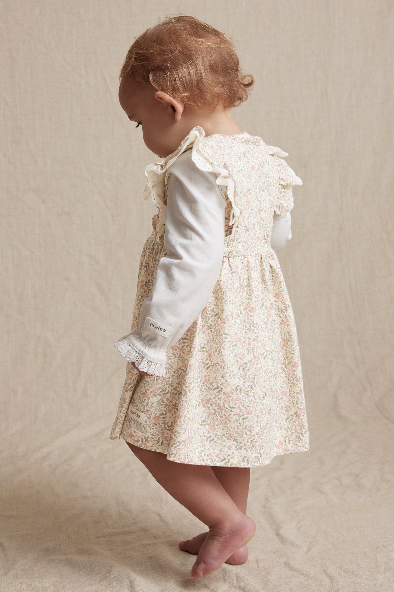 Cremeweißes geblümtes Jerseykleid mit Spitzenrüschen, von einem Baby über einem weißen Langarmshirt getragen.