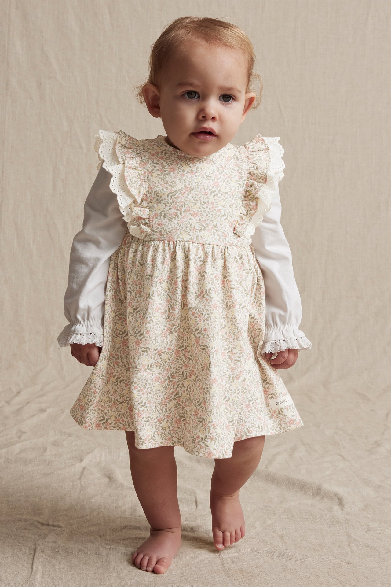 Vorderansicht eines Babys in einem cremefarbenen Blumenkleid mit Rüschen über einem weißen Langarmshirt.