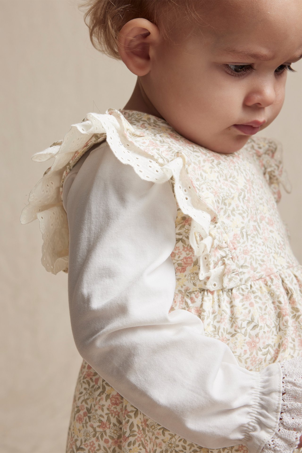 Seitenansicht Nahaufnahme eines Babys in cremefarbenem Blumenkleid mit Spitzenrüschen über weißem Langarmshirt.