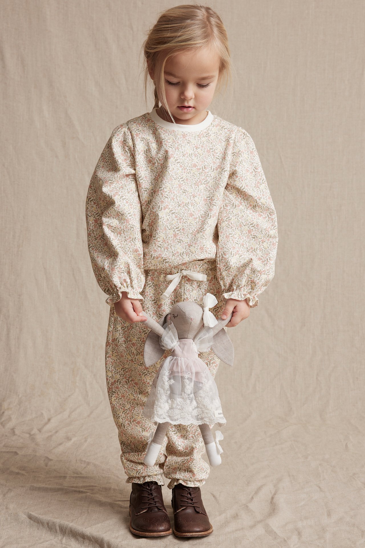 Front view: Kid in off-white floral cotton sweatshirt, matching trousers, brown boots, holding a doll.