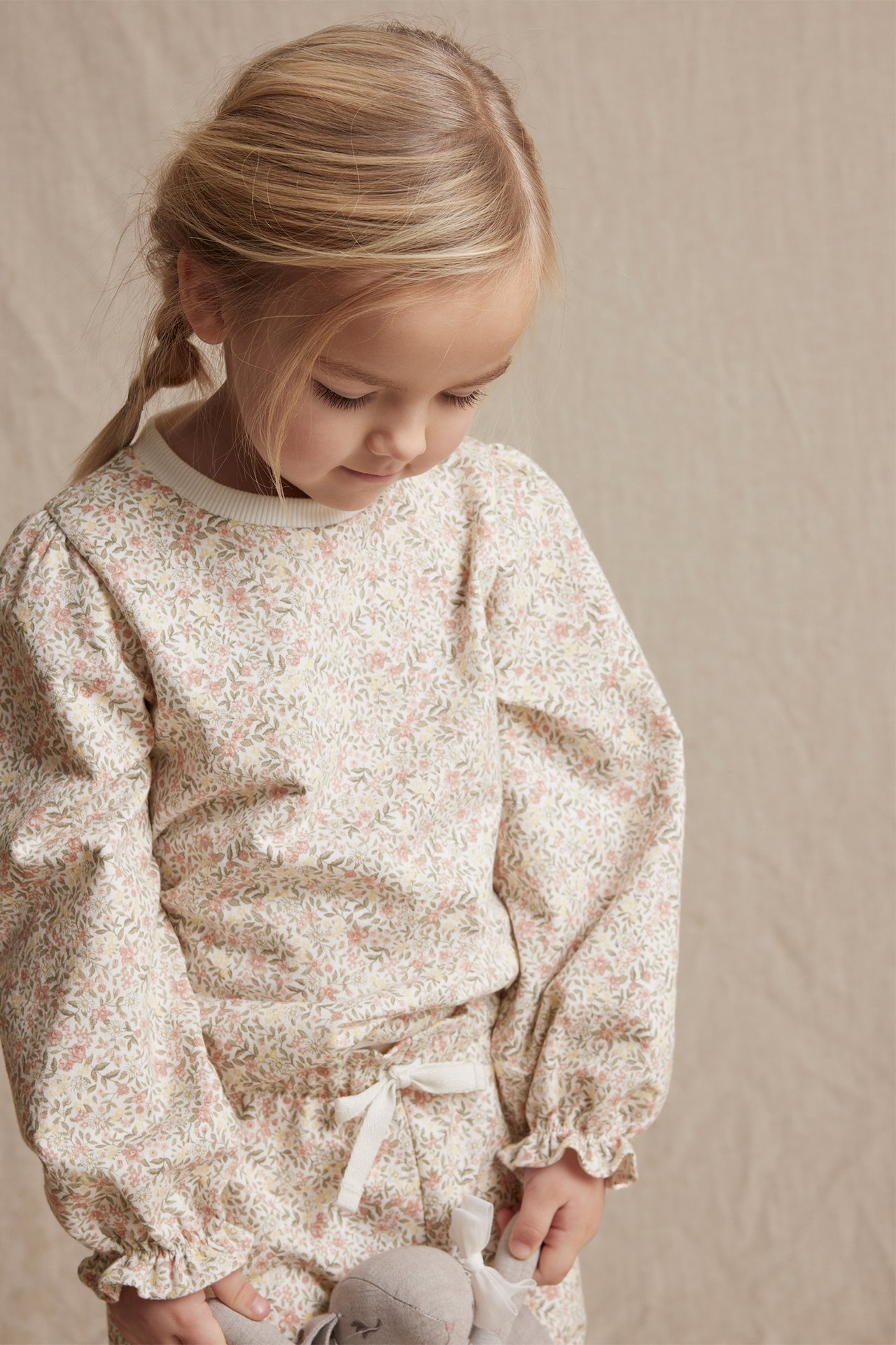 Front view: Kid in off-white floral cotton sweatshirt with puff sleeves, matching trousers, holding a grey toy.