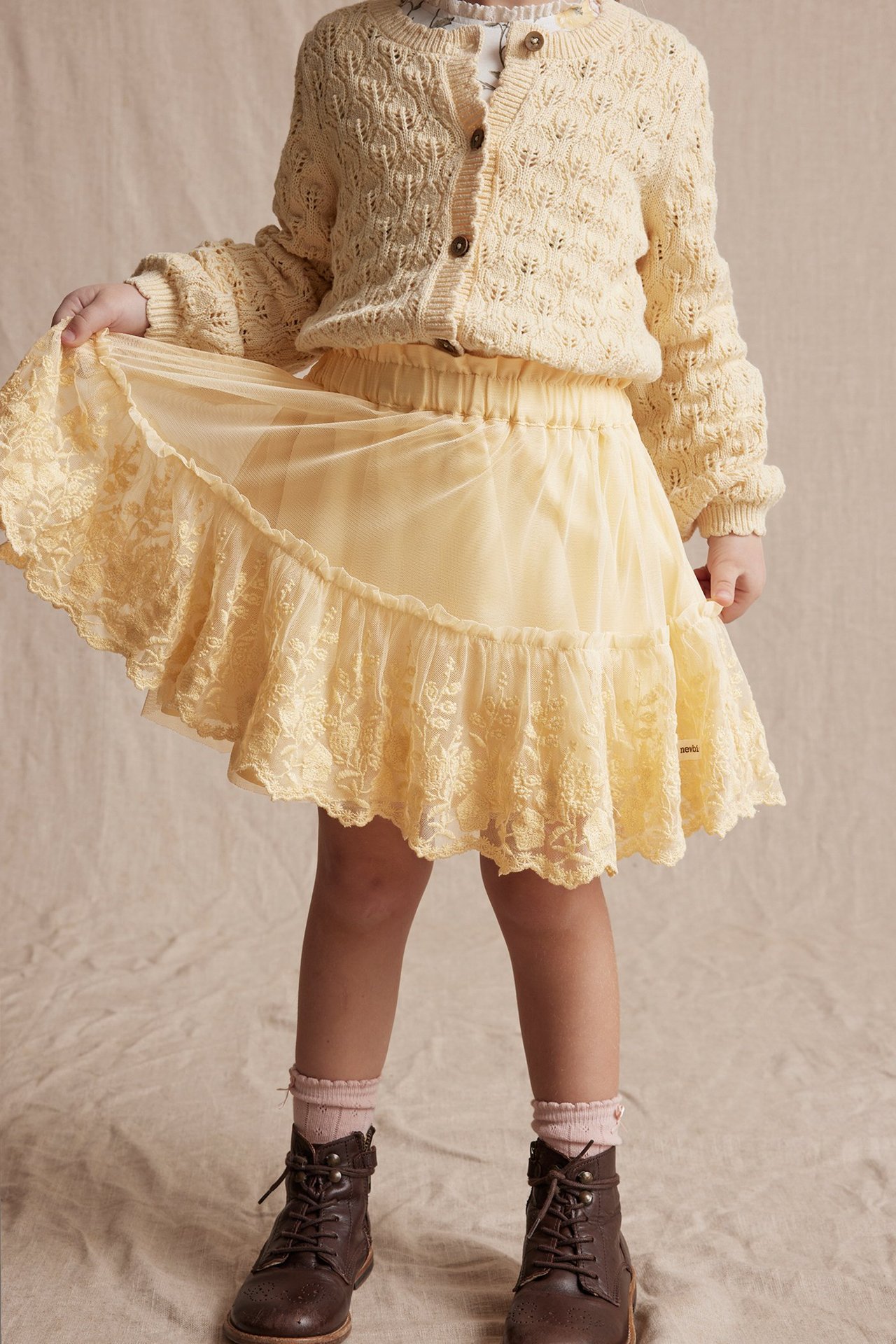 Front view: Kid in a yellow mesh skirt with embroidered ruffle, cream knitted cardigan, white top, and brown boots.