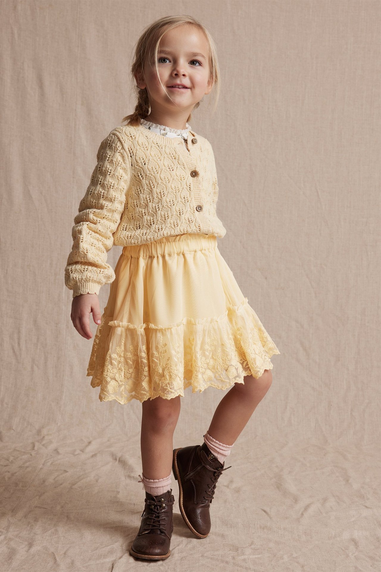 Front view of a kid in a yellow embroidered mesh skirt, yellow knitted cardigan, white blouse, and brown boots.