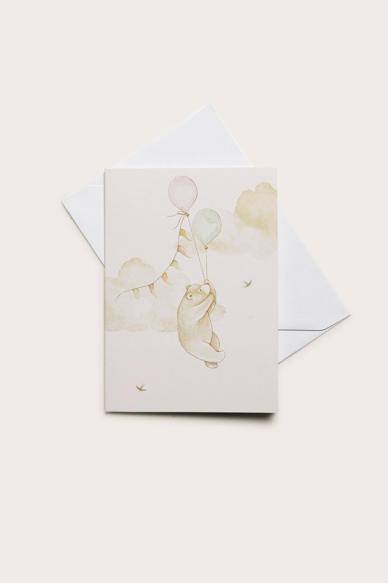 Draufsicht: Eine cremefarbene Klappkarte mit Teddybär und Ballons, mit einem weißen Umschlag.