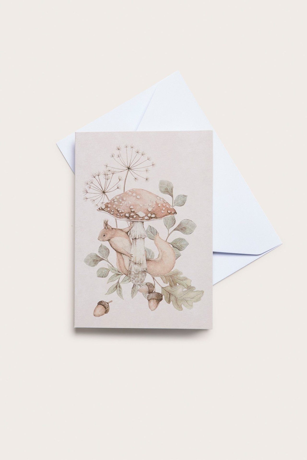 Draufsicht auf eine hellbeige Papierkarte mit Eichhörnchen-, Pilz- und Blattmuster, mit einem weißen Umschlag.