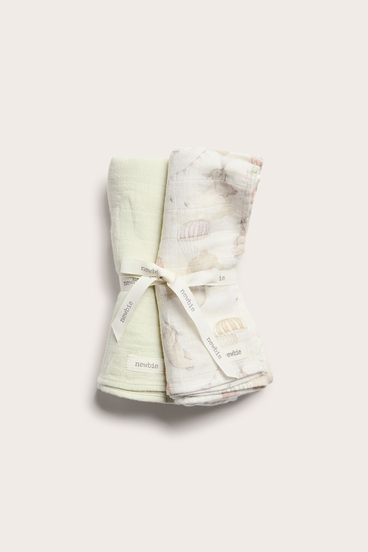 Front view: Two rolled muslin blankets, one offwhite patterned, one plain light green, tied with a ribbon.