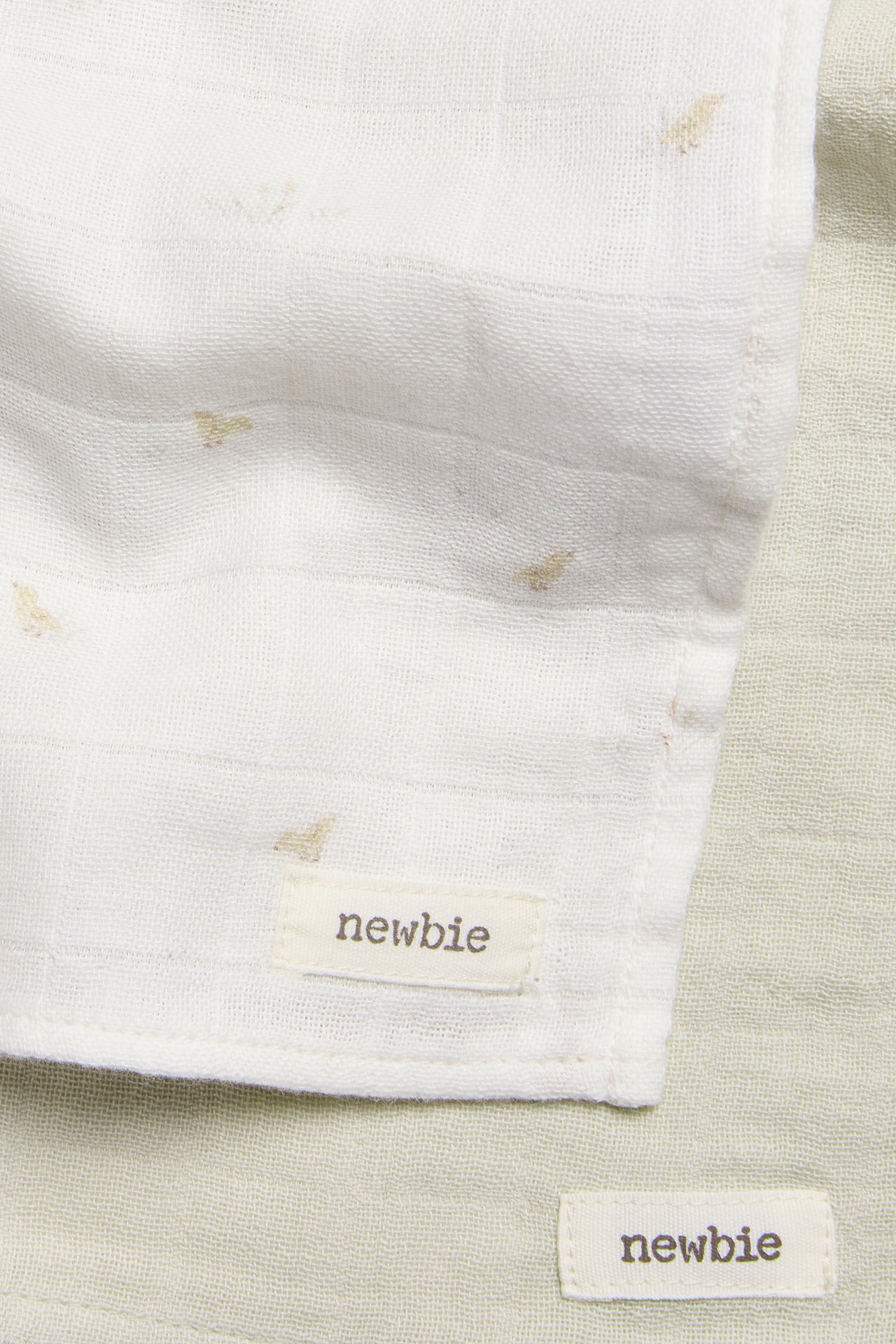 Close-up, top view of two cotton muslin blankets: one offwhite with small beige patterns, and one plain light green.