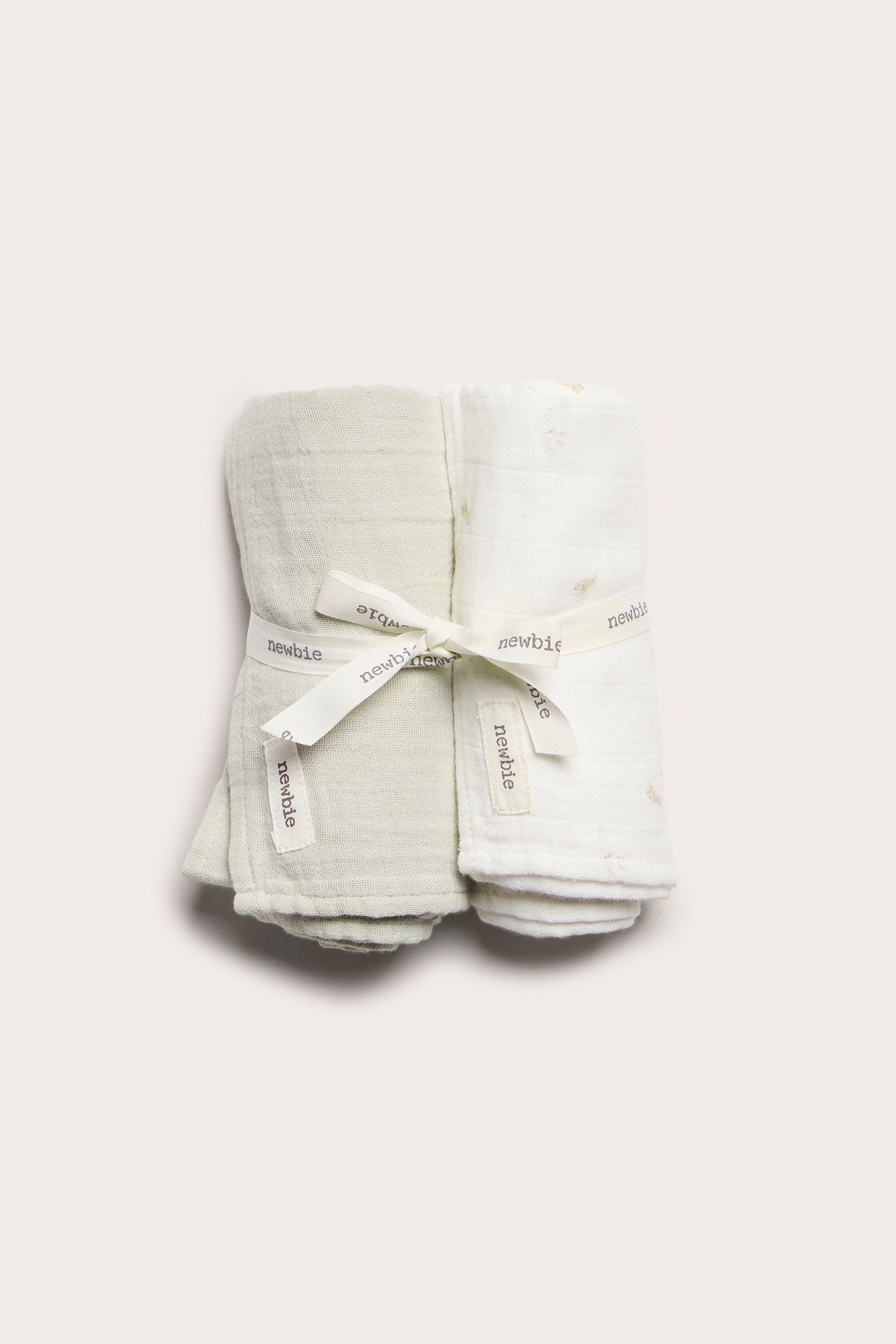 Top-down view of two rolled muslin blankets, one solid light green, one off-white patterned, tied with a ribbon.
