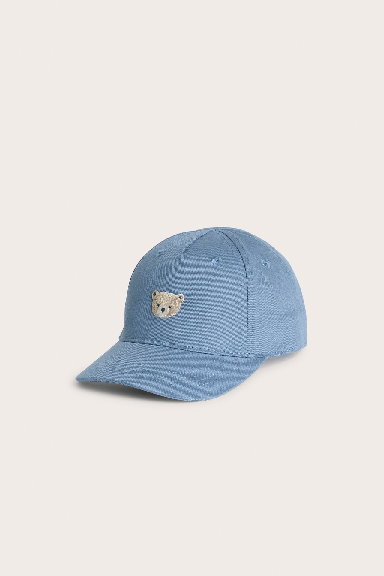 Schräge Vorderansicht einer hellblauen Baumwoll-Baby-/Kinderkappe mit kleinem gesticktem Bärengesicht.