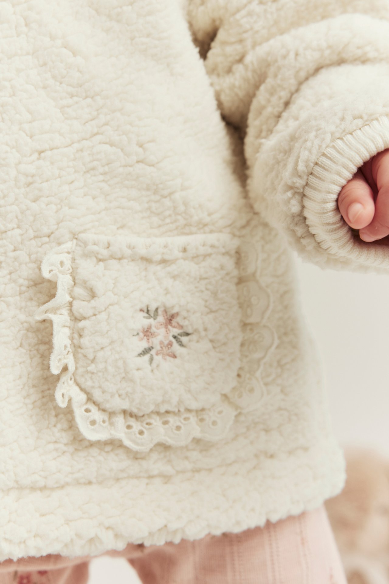 Nahaufnahme eines Babys in cremeweißer Piléjacke mit bestickter Tasche und Spitzenbesatz, dazu rosa Leggings.
