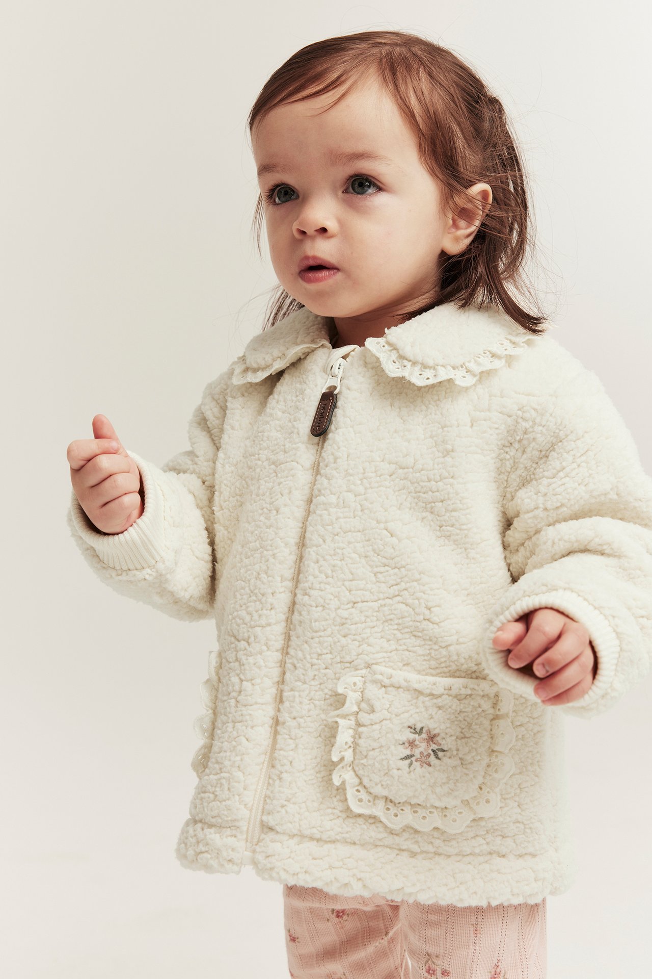 Vorderansicht eines Baby-Mädchens in cremeweißer Piléjacke mit Spitzenkragen, bestickten Taschen und rosa gemusterter Hose.