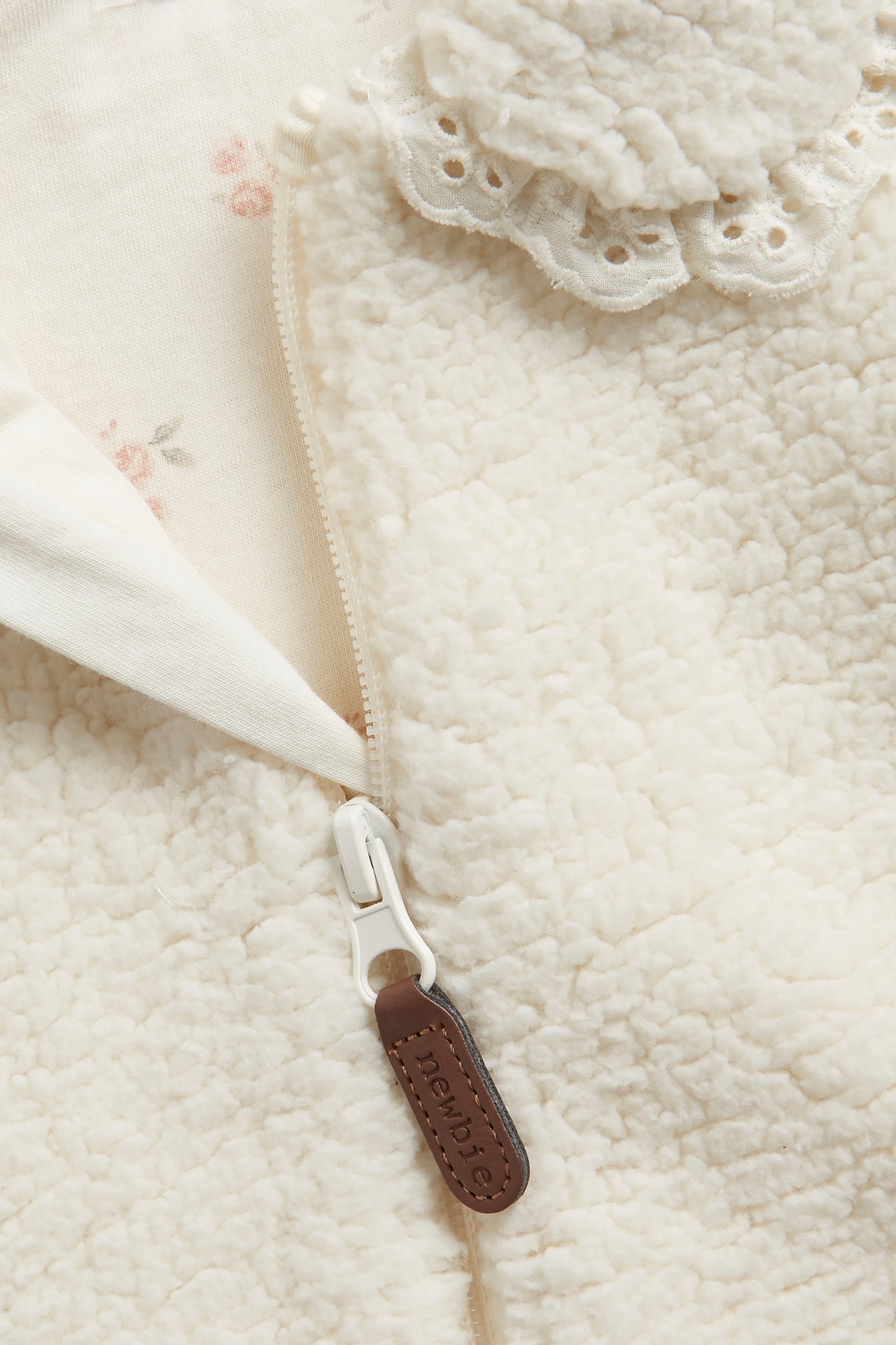 Nahaufnahme einer cremeweißen Baby-Fleecejacke mit weißem Reißverschluss, Spitzenkragen und geblümtem Baumwollfutter.