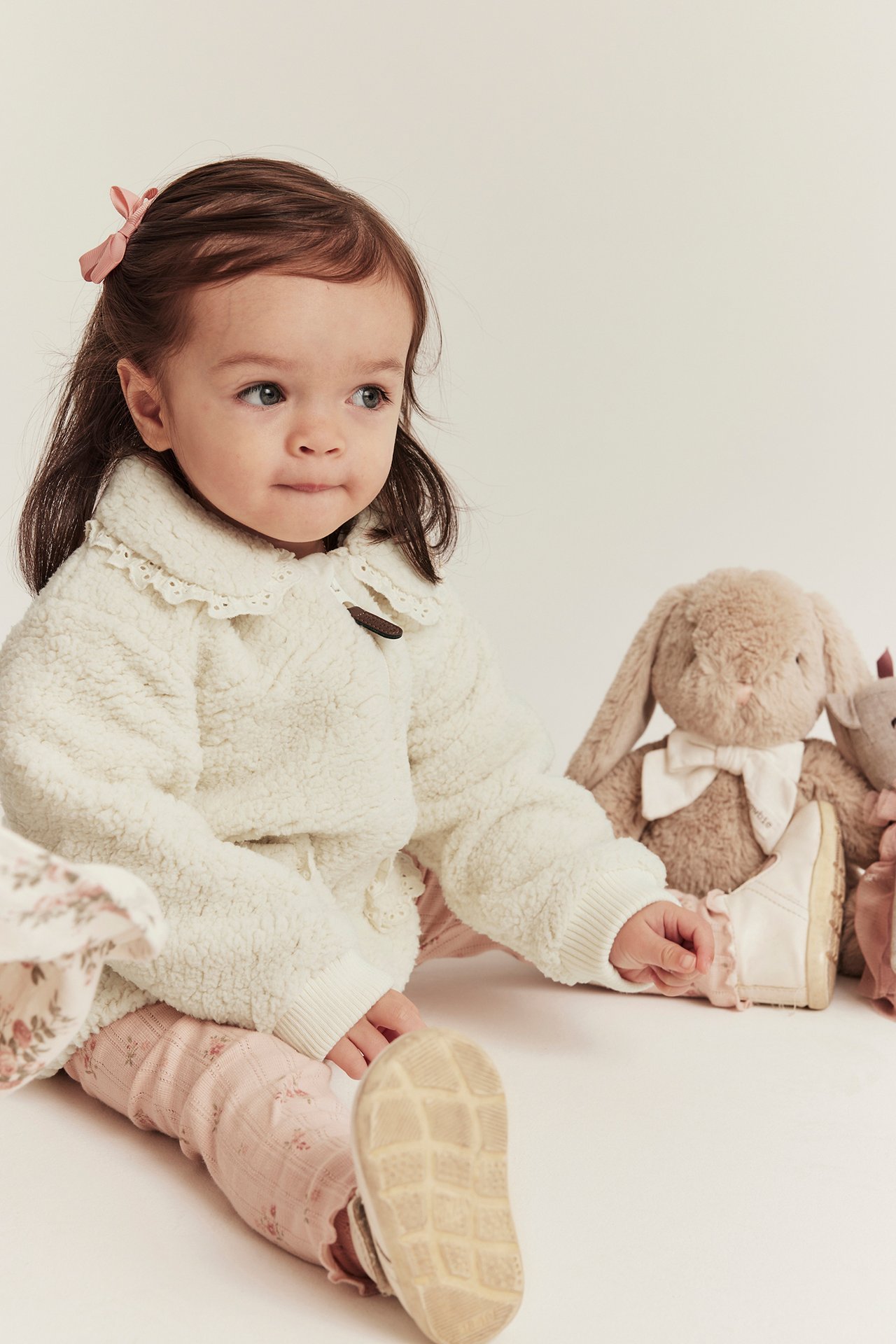 Vorderansicht eines Babys in off-white Pile-Jacke mit Spitzenkragen, rosa Blumen-Leggings und weißen Schuhen.