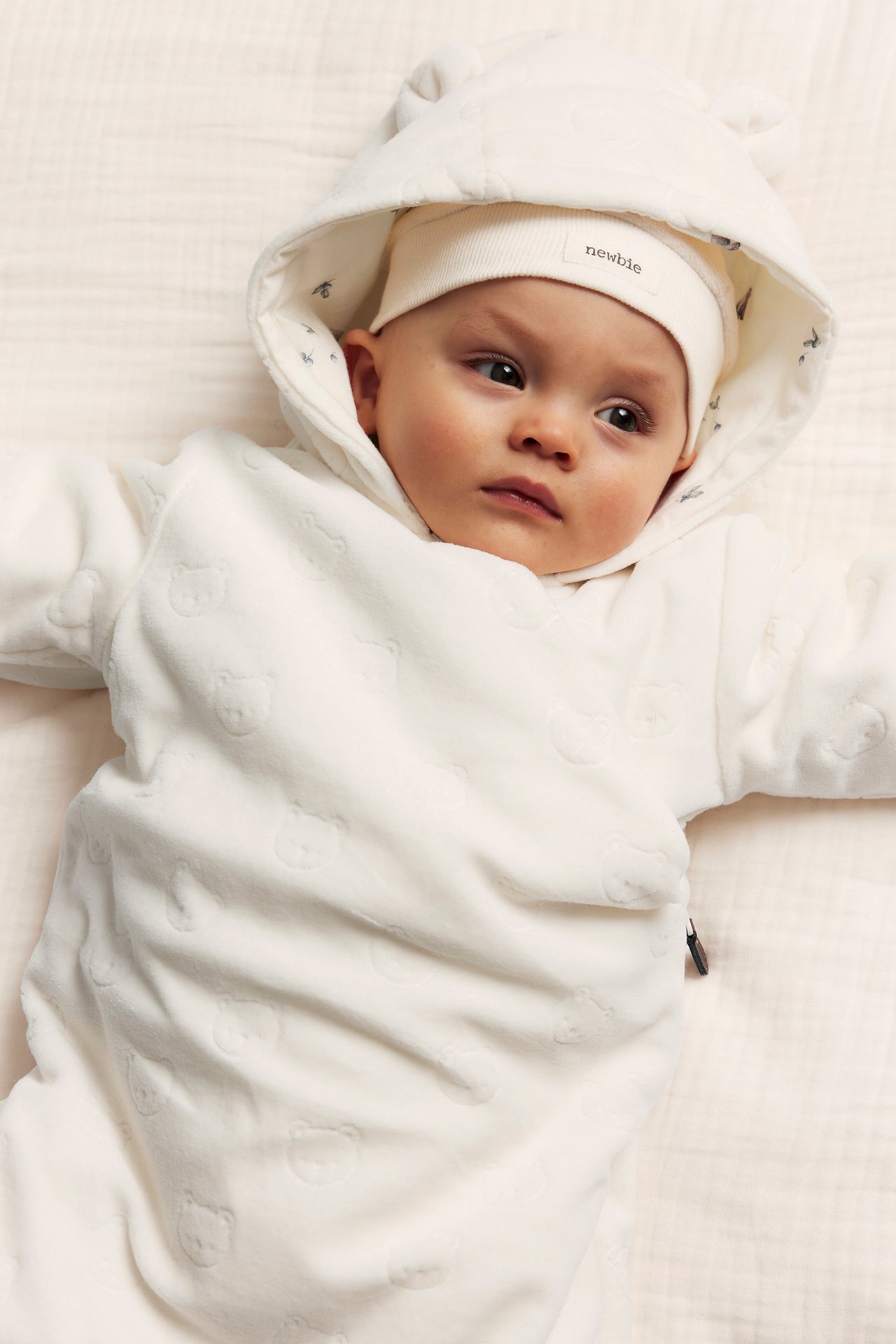 Close-up of a baby in an off-white velour coverall with embossed bears, ear hood, and a beanie.