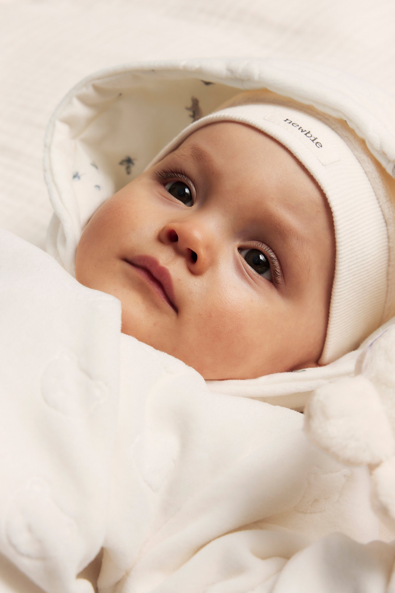 Close-up of a baby wearing an off-white velour coverall with an embossed pattern and a white beanie.