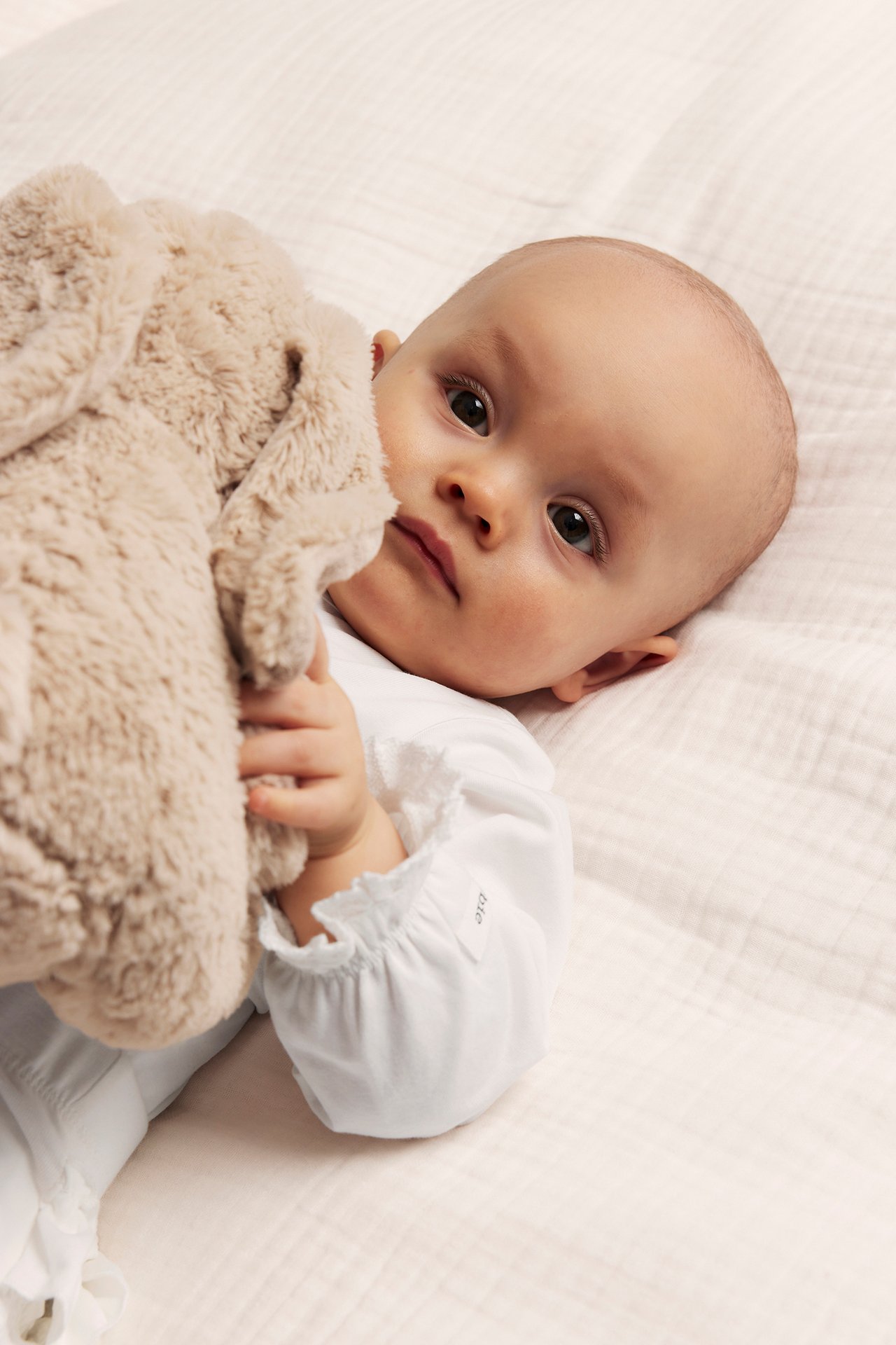 Close-up of a baby in an off-white velour coverall, holding a beige plush toy.