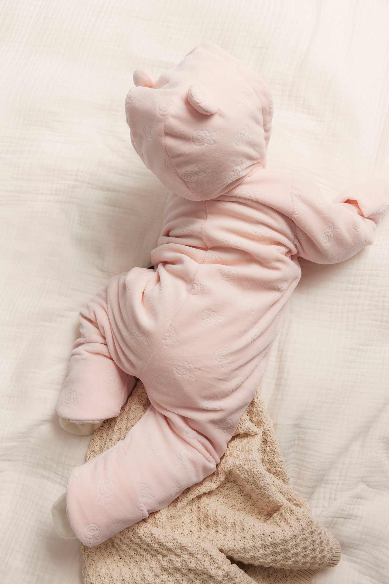 Draufsicht auf Baby in rosa Velours-Overall mit Prägemuster und Kapuze mit Ohren, auf Decke liegend.