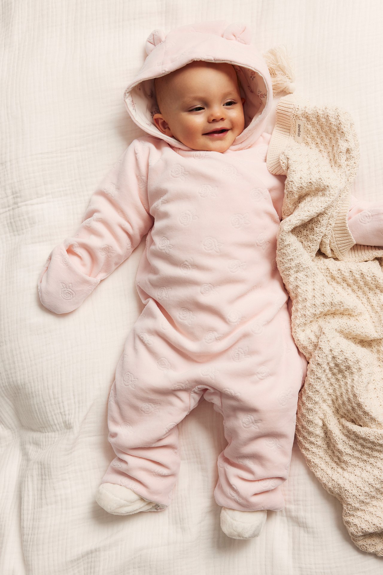 Baby in rosa Velour-Overall mit geprägtem Rosenmuster und Ohrenkapuze, neben beiger Strickdecke.