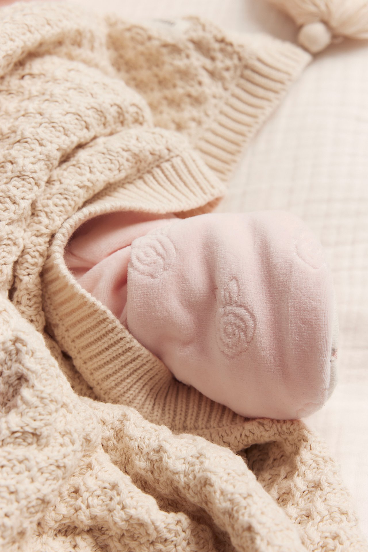 Nahaufnahme eines Babyfußes in rosa Velours-Overall mit Prägemuster, unter einer beigen Strickdecke.