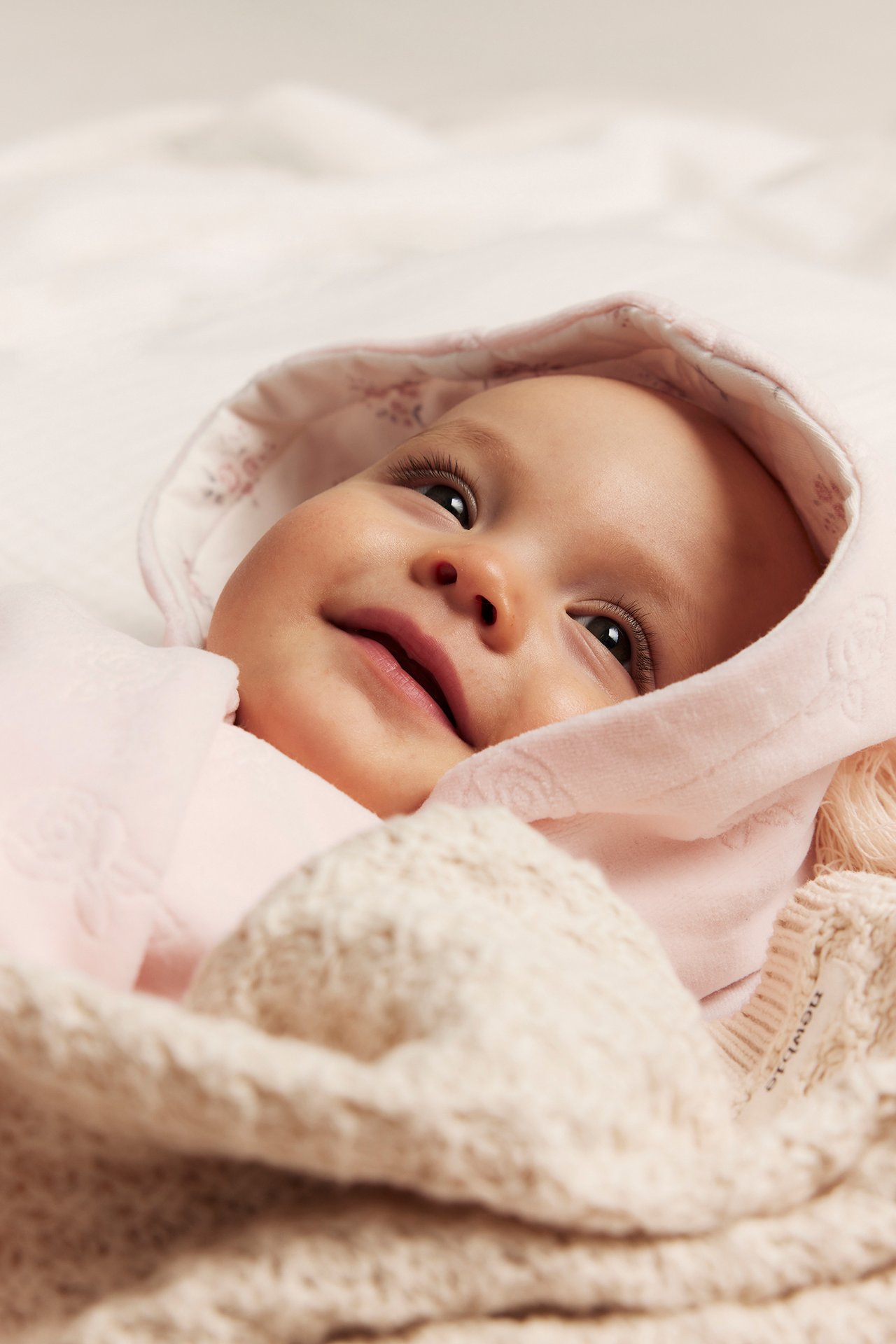 Nahaufnahme eines Babys in einem rosa Velours-Overall mit Prägemuster und bedruckter Kapuze, unter einer Strickdecke.