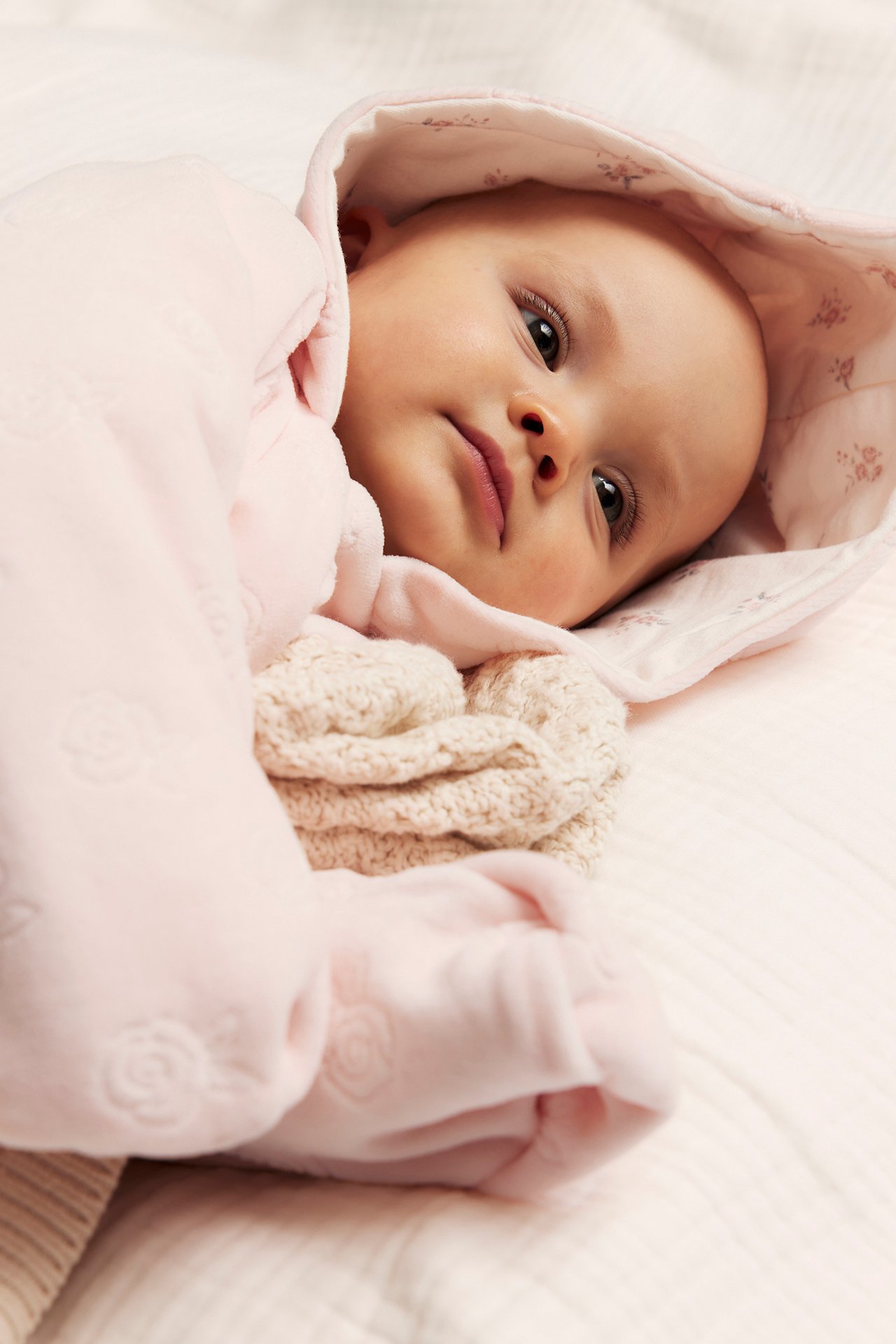 Nahaufnahme eines Babys in einem rosa Velours-Overall mit Prägemuster und floral gefütterter Kapuze.