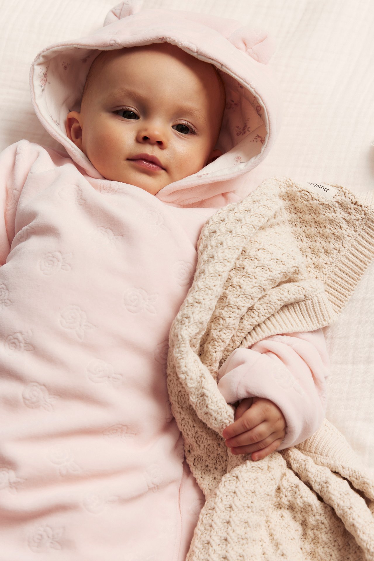 Draufsicht: Baby in rosa Velours-Overall mit geprägten Rosen, Ohrenkapuze, hält eine beige Strickdecke.