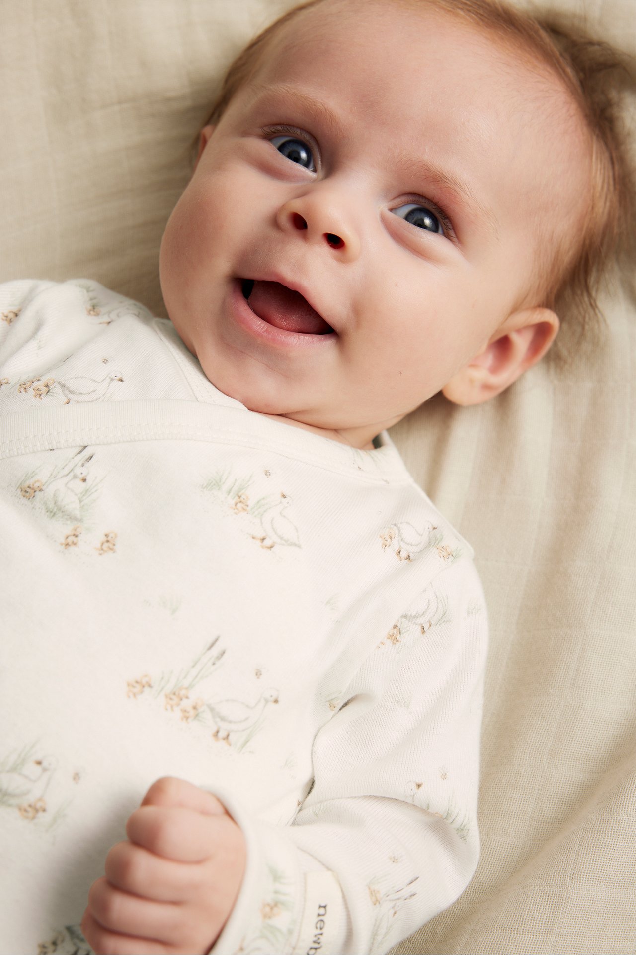 Nahaufnahme eines lächelnden Babys in einem cremeweißen gerippten Wickelbody mit Entenmuster.