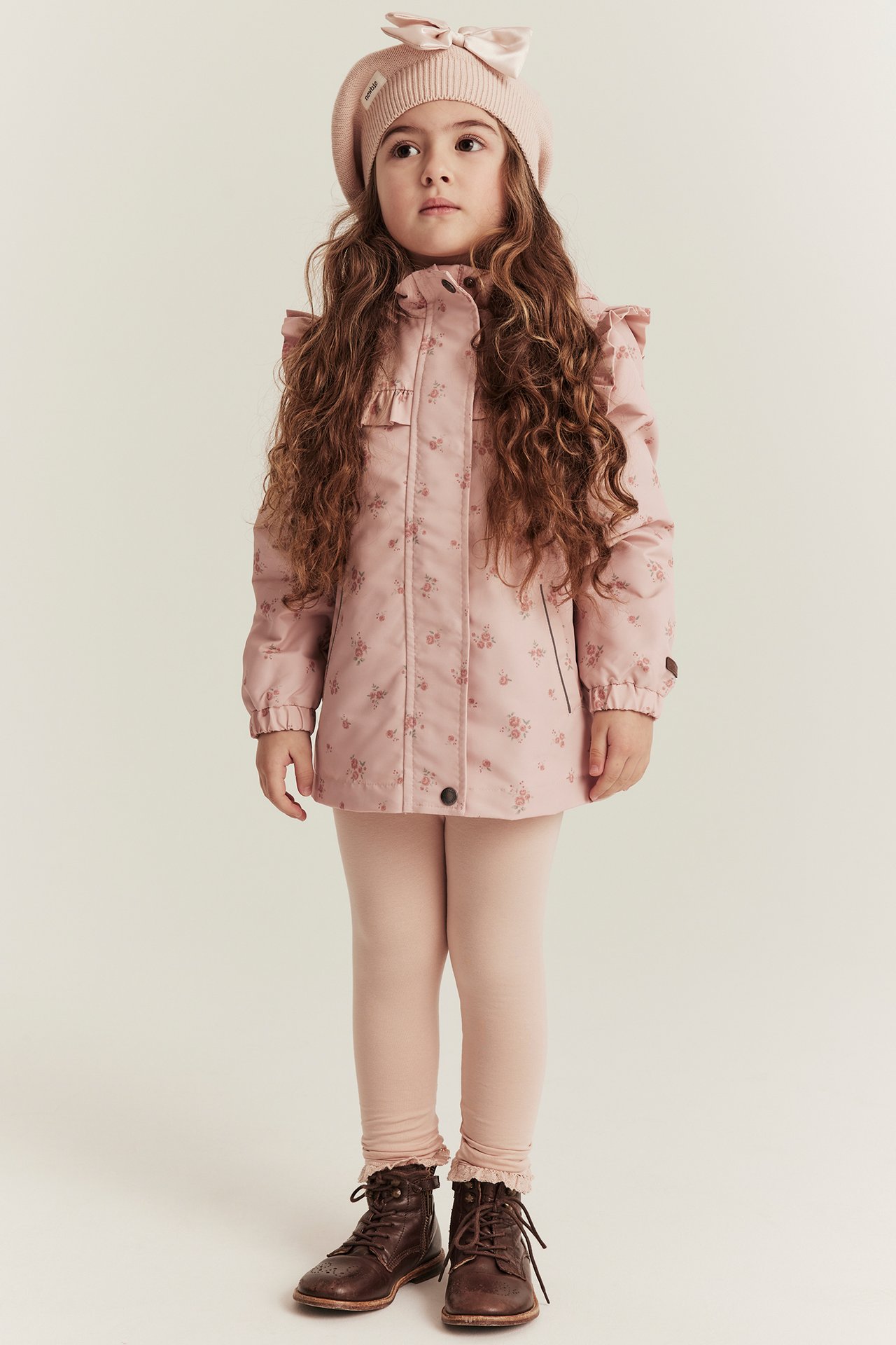 Kind, Vorderansicht: hellrosa Blumenjacke mit Rüschen, rosa Baskenmütze, hellrosa Leggings und braune Stiefel.