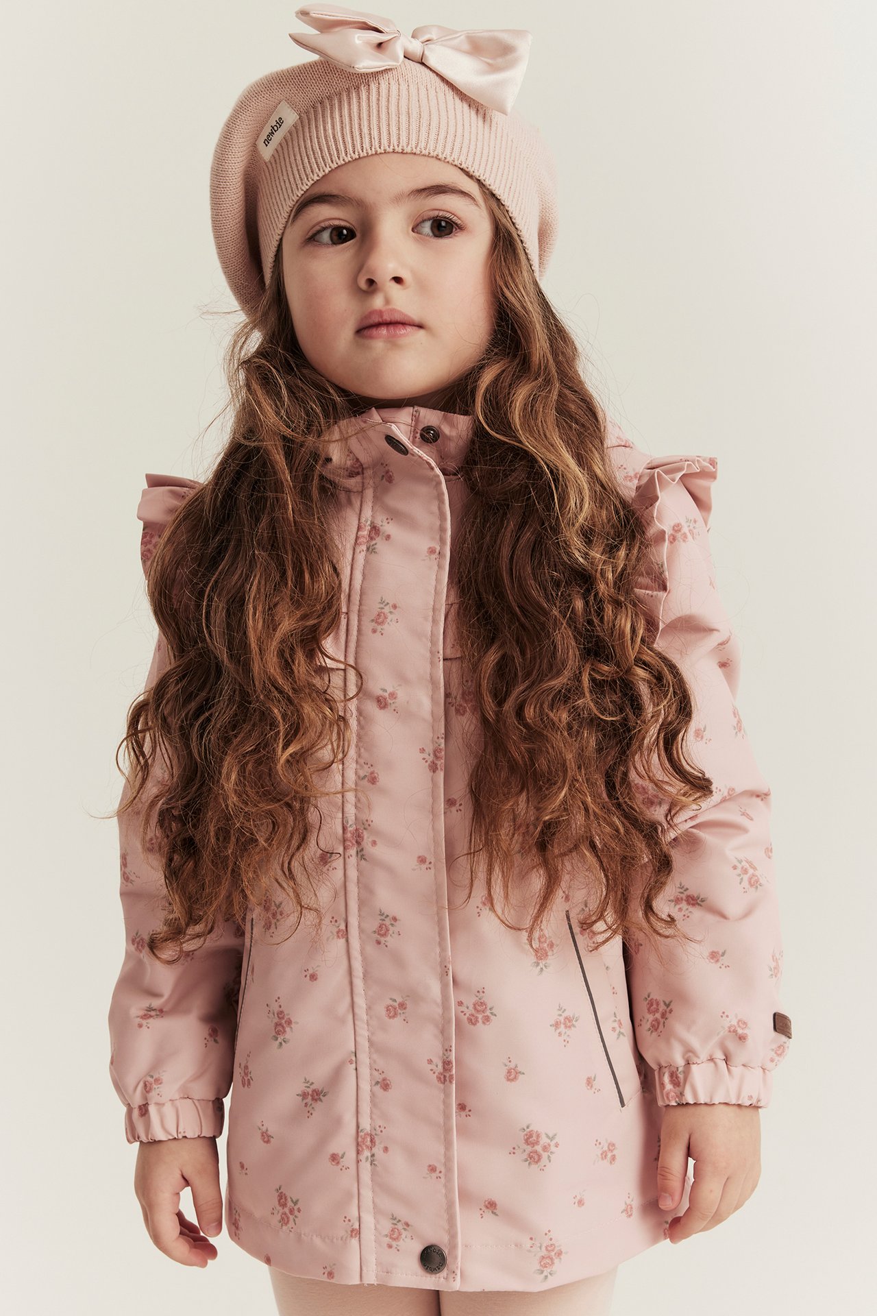 Vorderansicht eines Kindes in hellrosa Blumenjacke mit Rüschen und hellrosa Strickbarett mit Schleife.