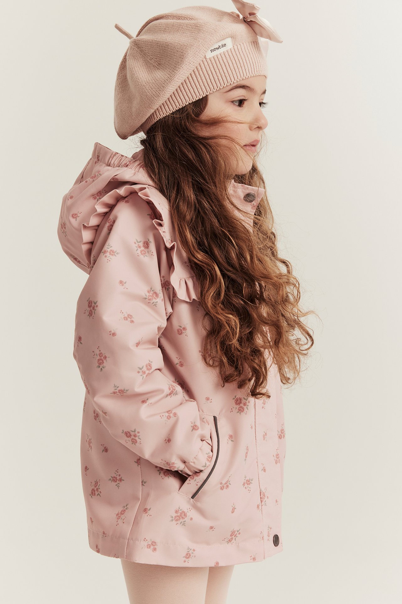 Seitenansicht eines Kindes in einer hellrosa geblümten wasserabweisenden Jacke mit Rüschen und einer rosa Strickmütze.