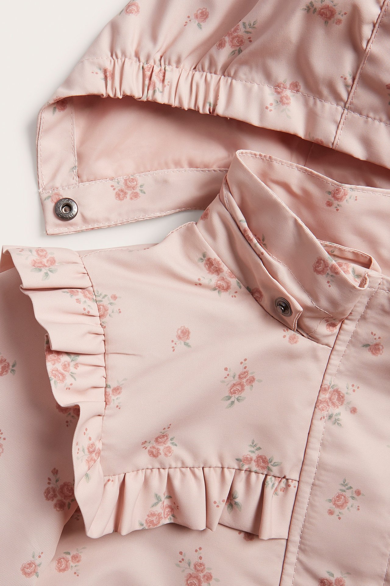 Nahaufnahme einer hellrosa Kinderjacke mit Blumenmuster, Rüschen und abnehmbarer Kapuze.