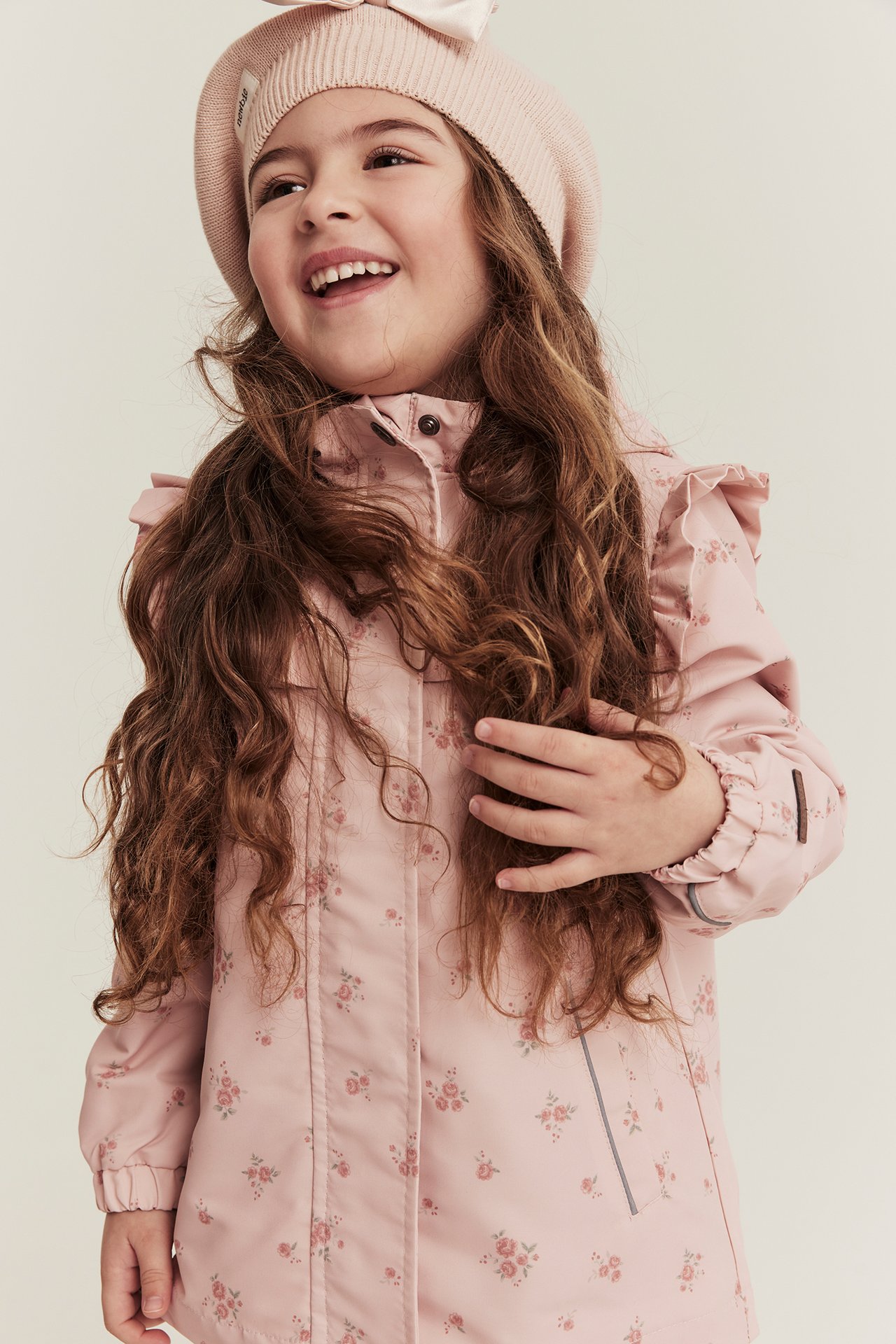 Vorderansicht eines Kindes in einer hellrosa Blumenjacke mit Rüschen und einer hellrosa Strickmütze.