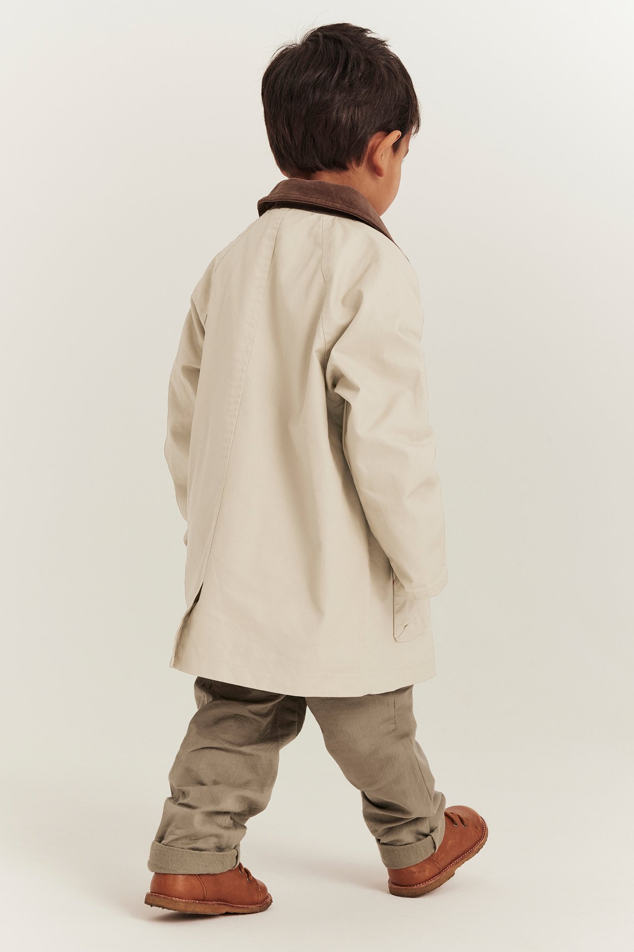Rückenansicht eines Kindes in beiger Baumwolljacke mit braunem Cordkragen, khakifarbenen Hosen und braunen Schuhen.