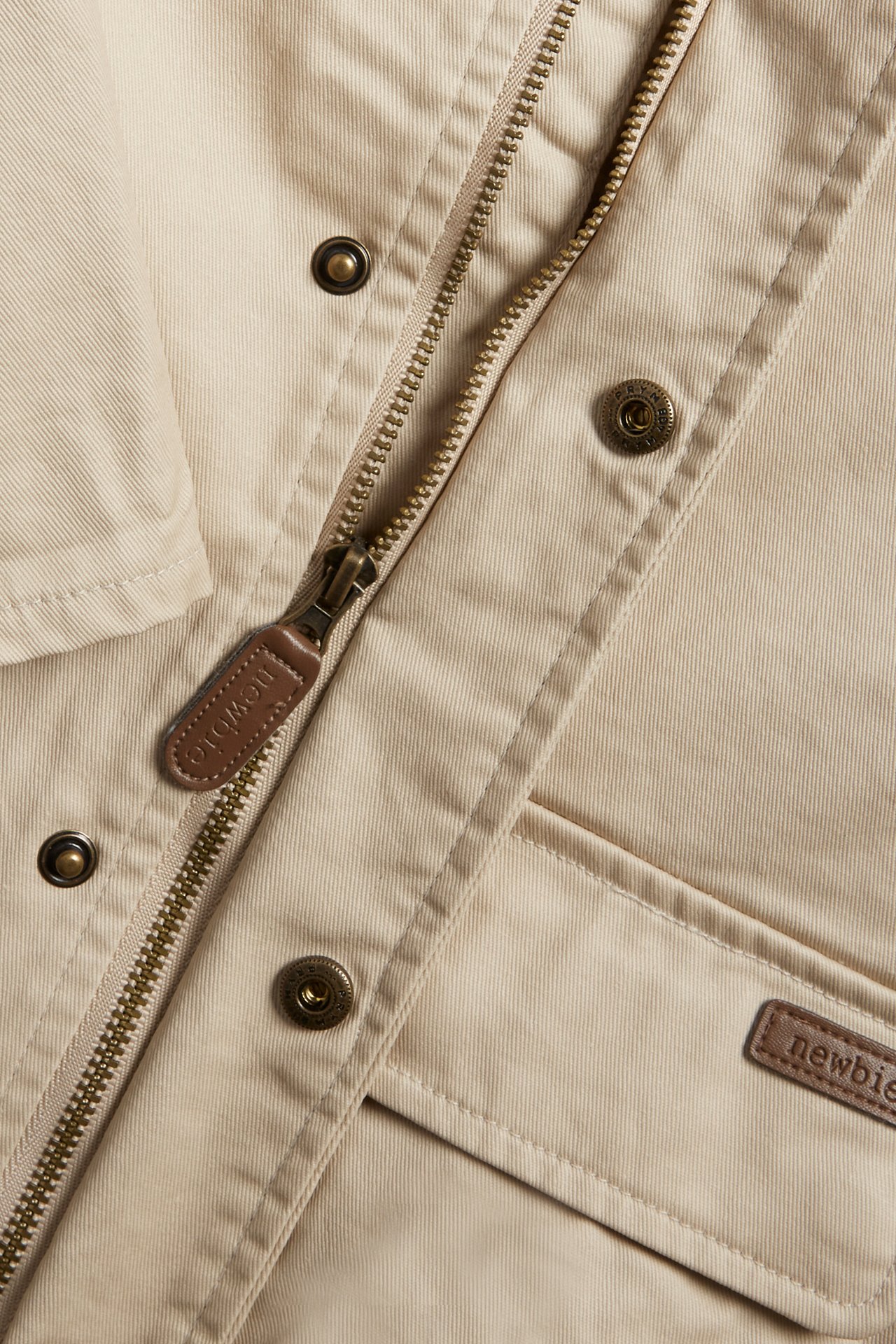Nahaufnahme von vorne einer beigen Baumwolljacke mit Reißverschluss, Druckknöpfen und braunem Markenetikett.