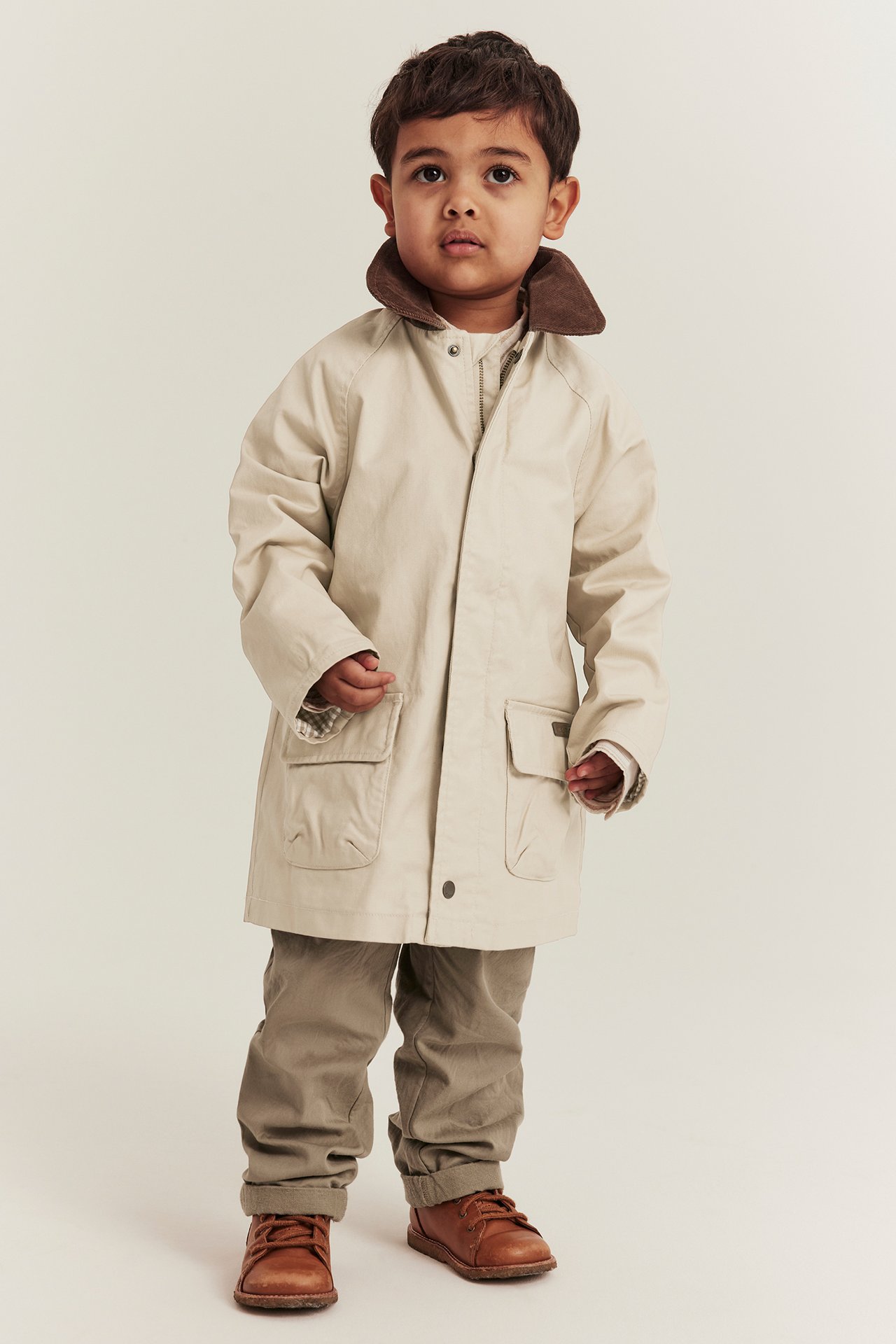 Vorderansicht: Kind in beiger Baumwolljacke mit braunem Cordkragen, beiger Hose und braunen Schuhen.