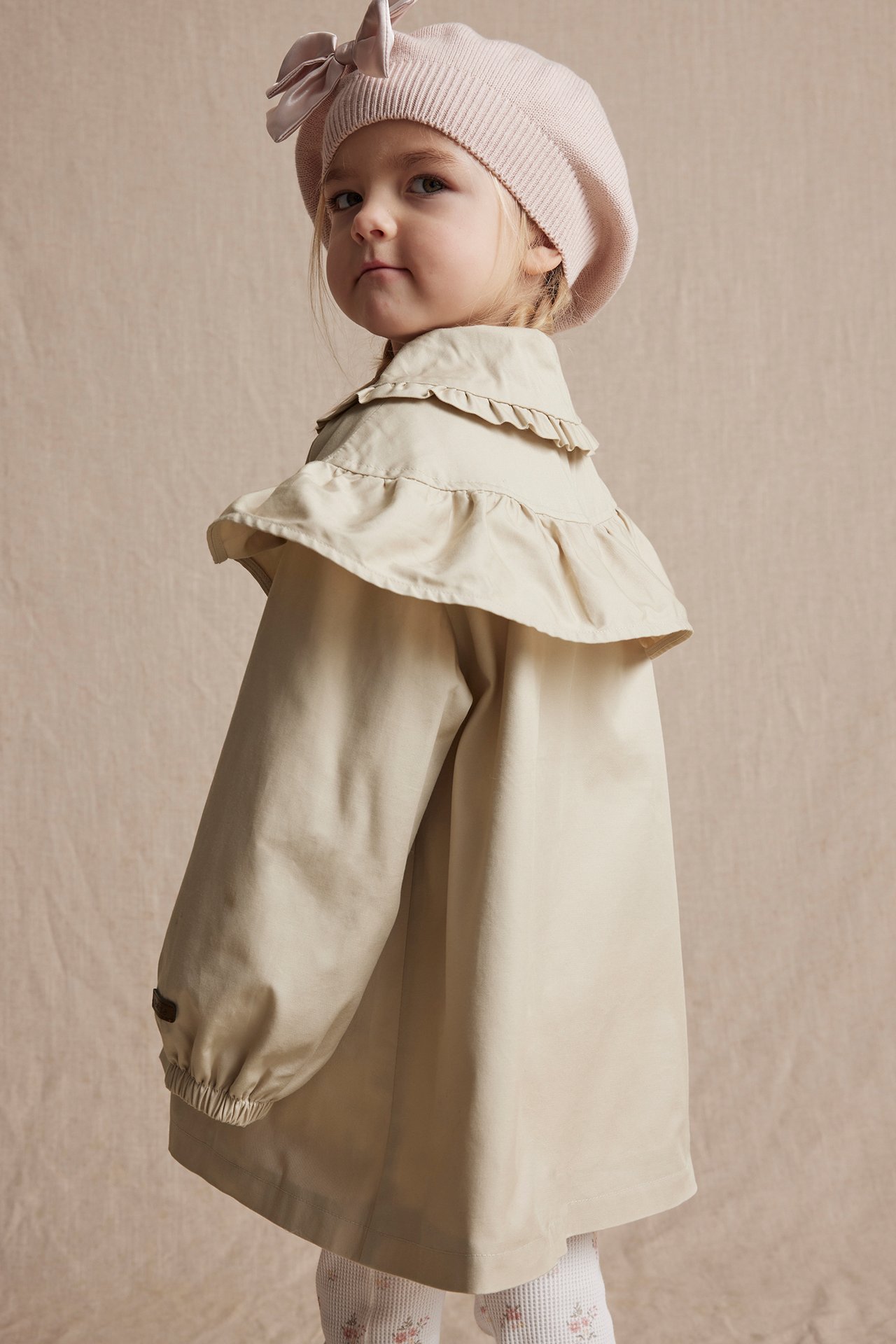 Side view of a kid wearing a beige cotton jacket with ruffles, a pink knitted beret, and white floral leggings.