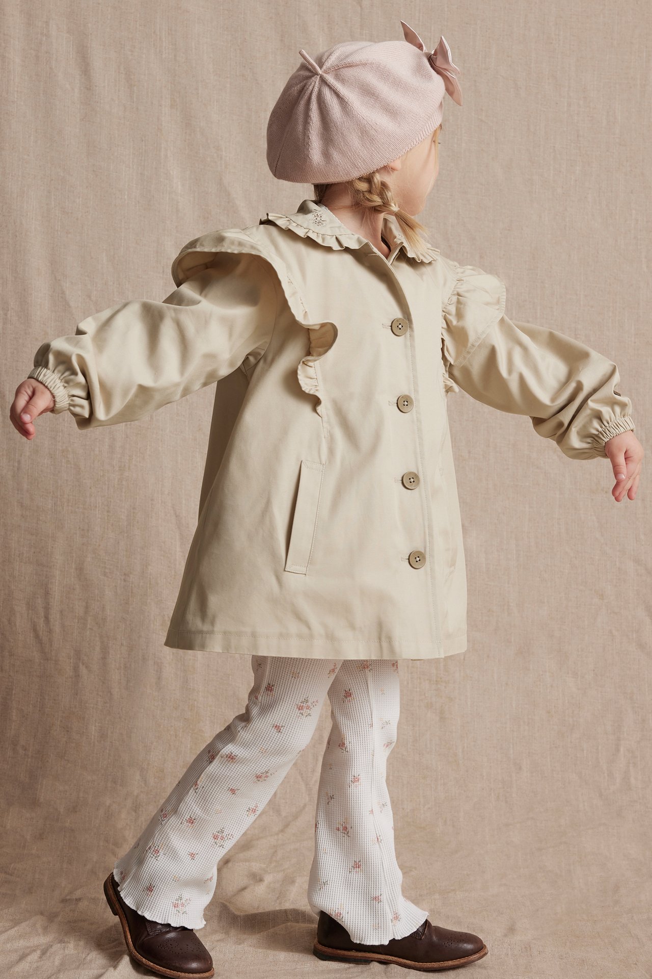 Back-side view of a kid wearing a beige ruffled cotton jacket, white floral leggings, a pink beret, and brown shoes.