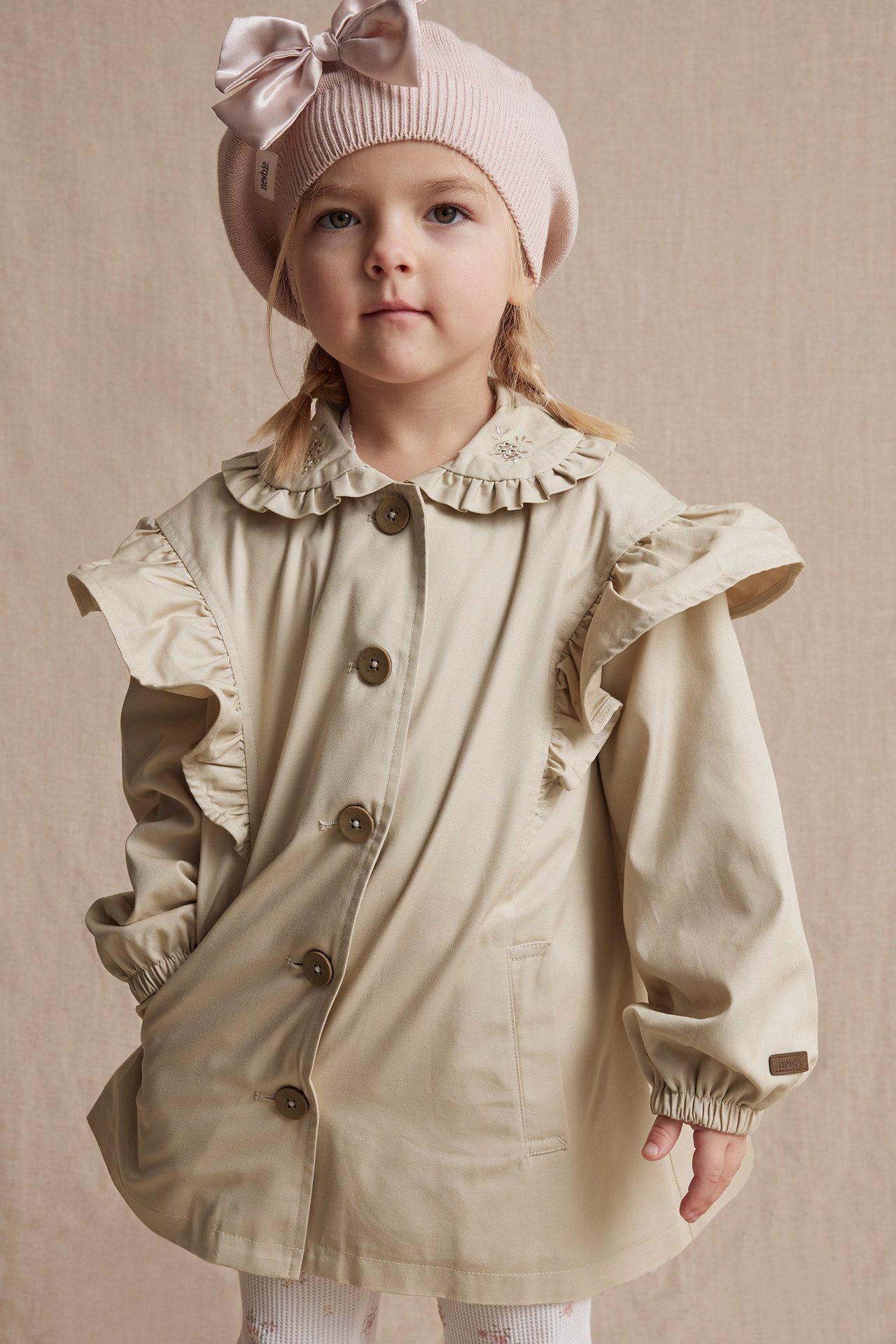 Front view of a kid in a beige ruffled, embroidered jacket and a pink bow beret.