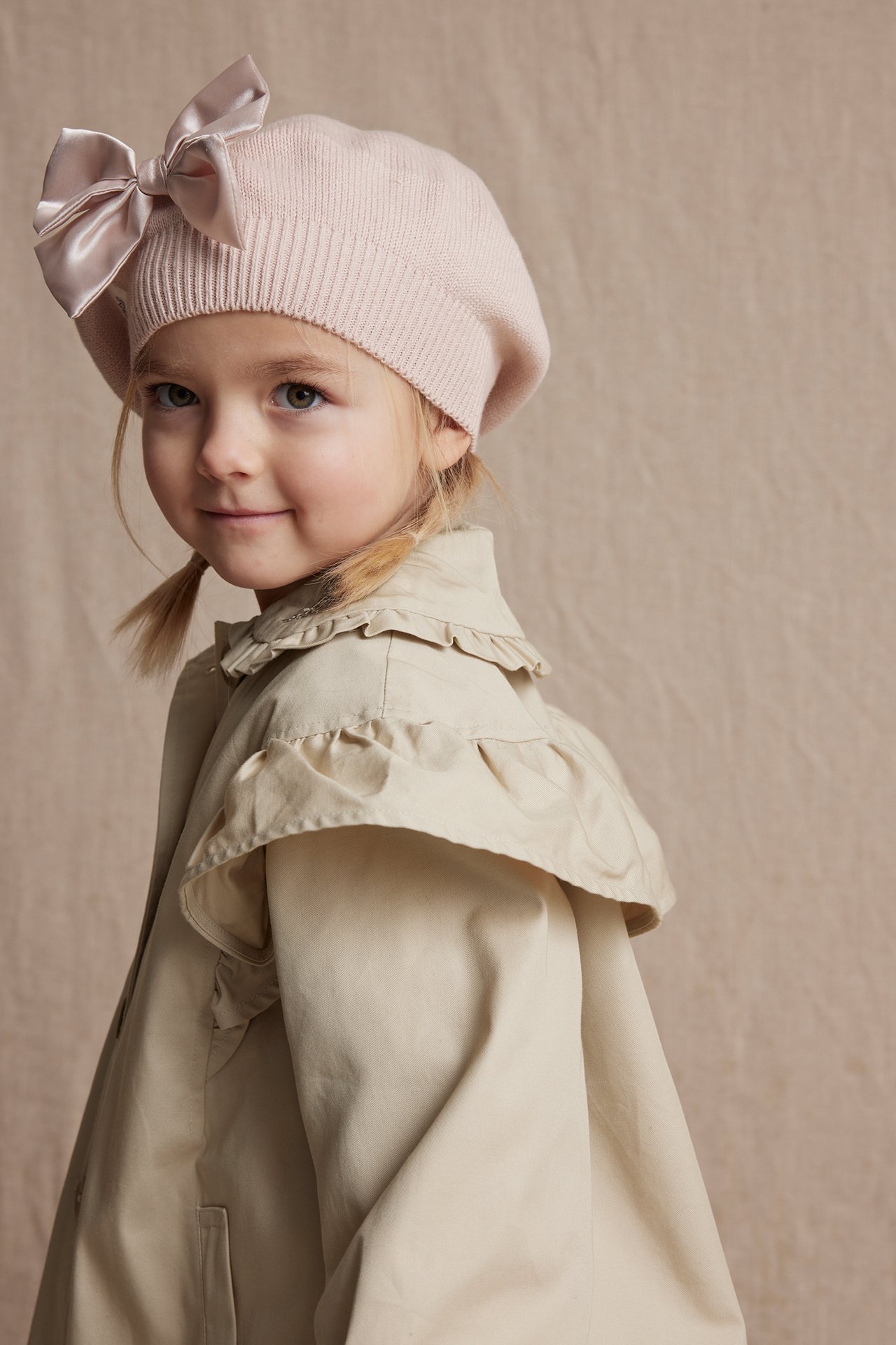 Side-front view of a kid wearing a beige cotton jacket with ruffles and a pink knitted beret with a bow.