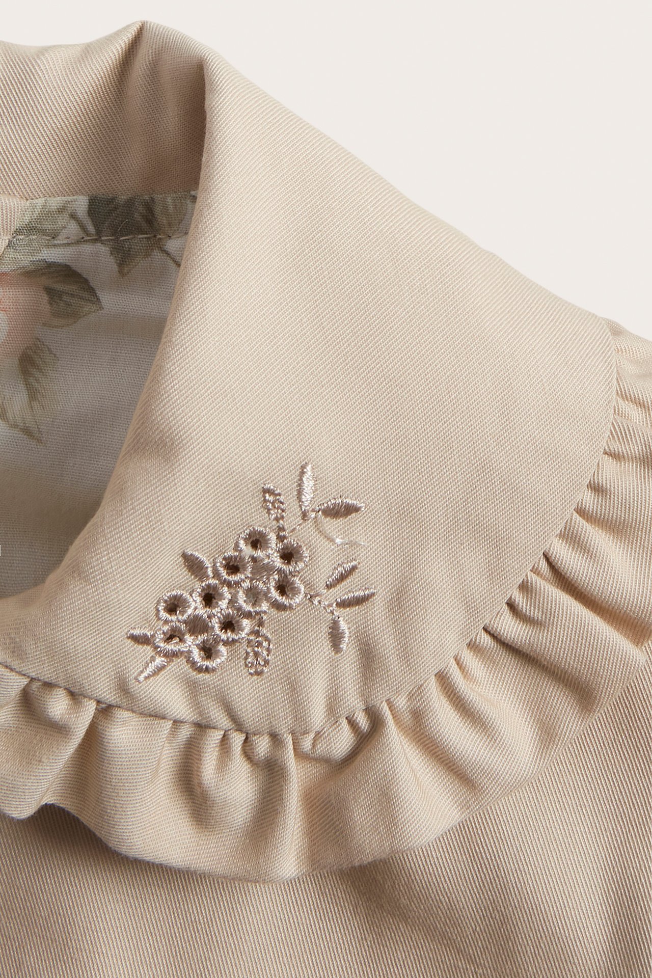 Close-up of a beige cotton jacket collar with ruffle trim, floral embroidery, and a patterned lining.