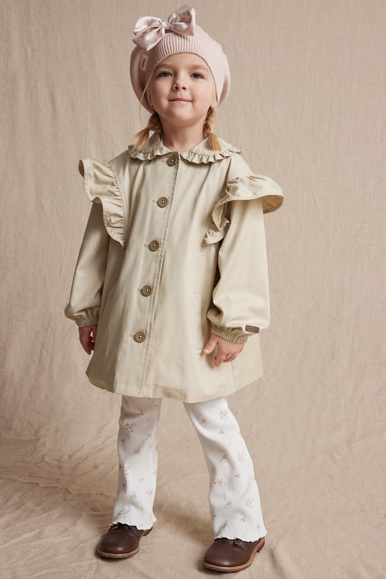 Front view of a kid in a beige ruffled cotton jacket, white floral pants, brown shoes, and a pink beret.