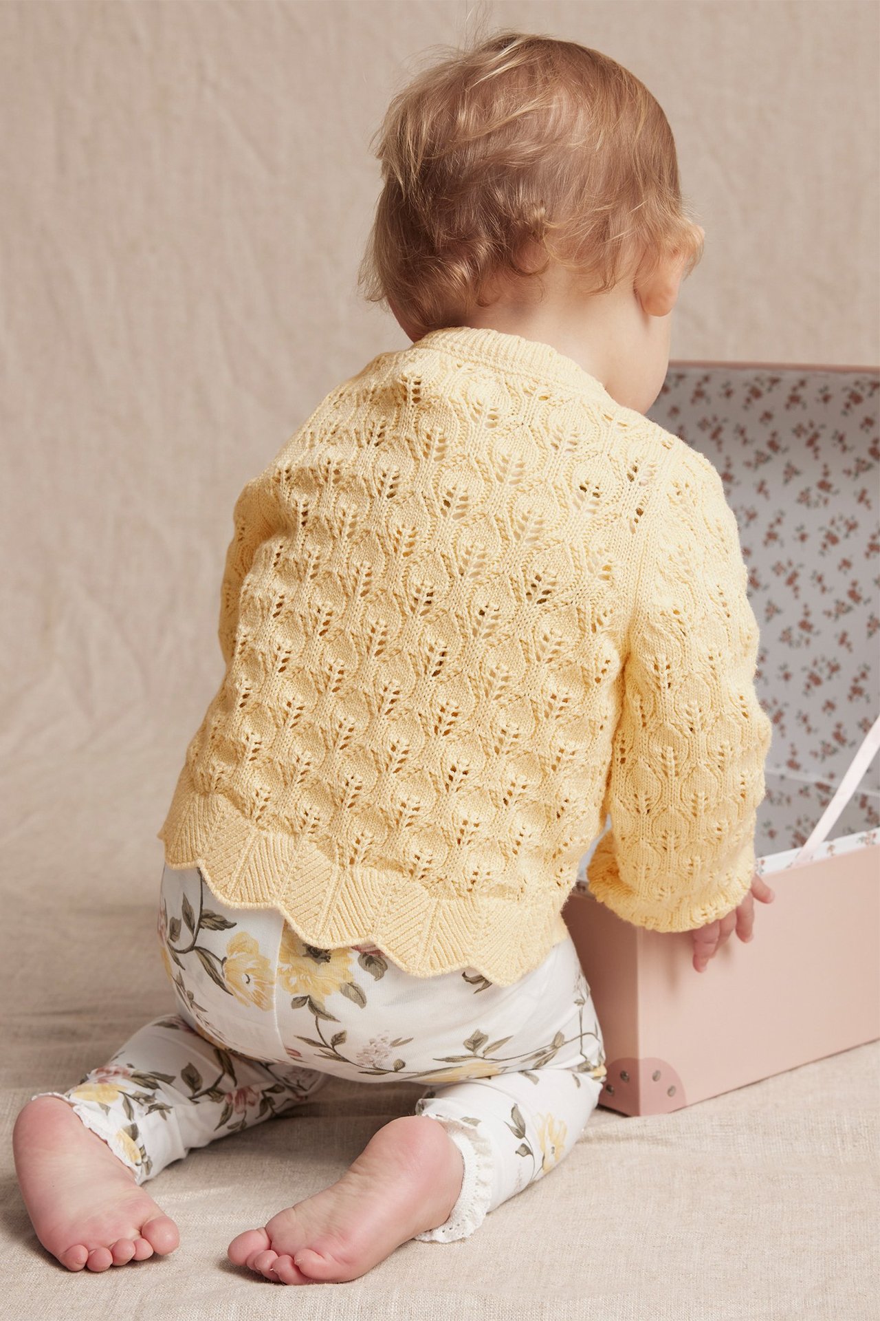 Back view of a baby wearing a yellow pattern-knitted cotton cardigan with scalloped hem and white floral leggings.