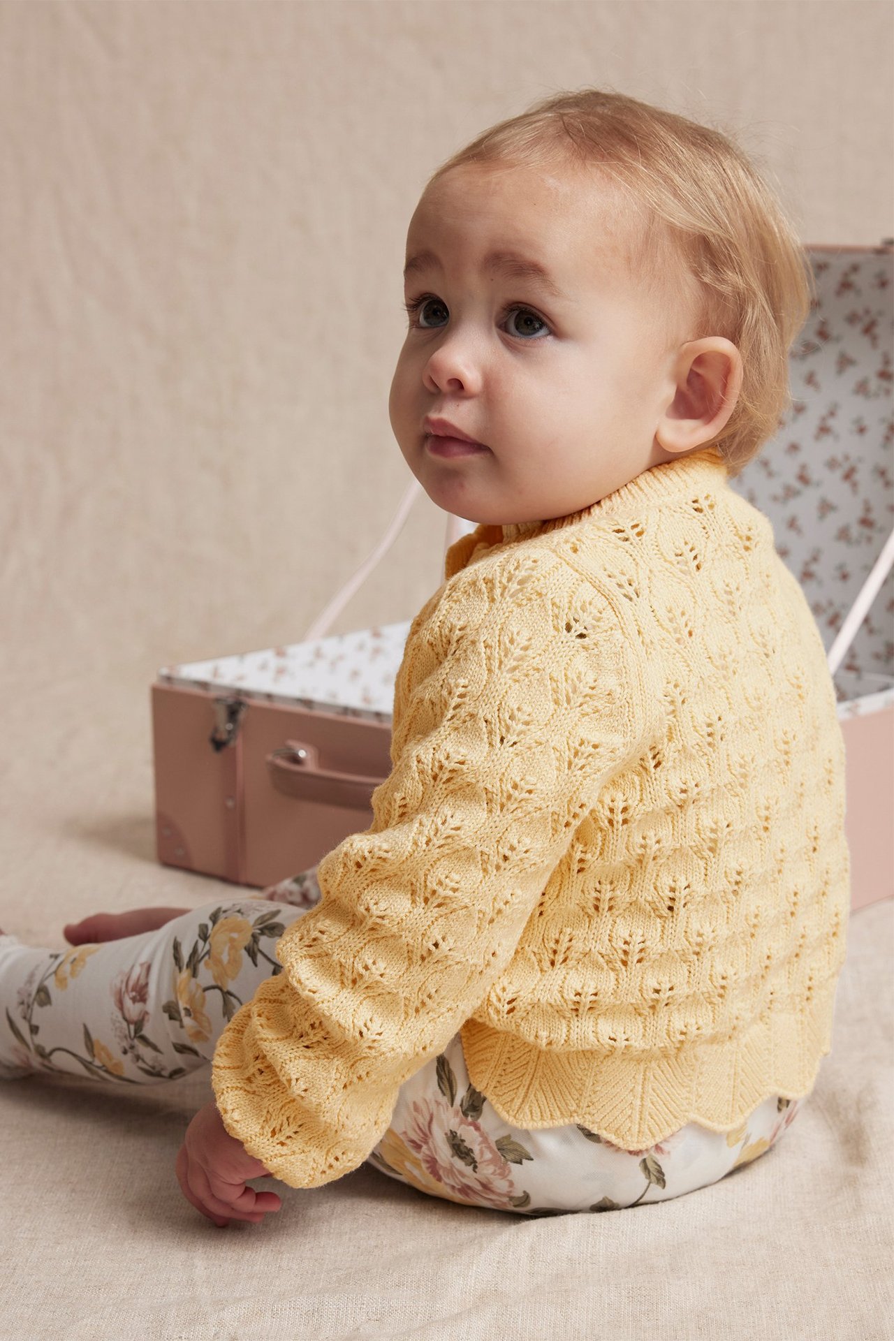 Side view of a baby wearing a yellow patterned knit cardigan with scalloped edges over floral trousers.