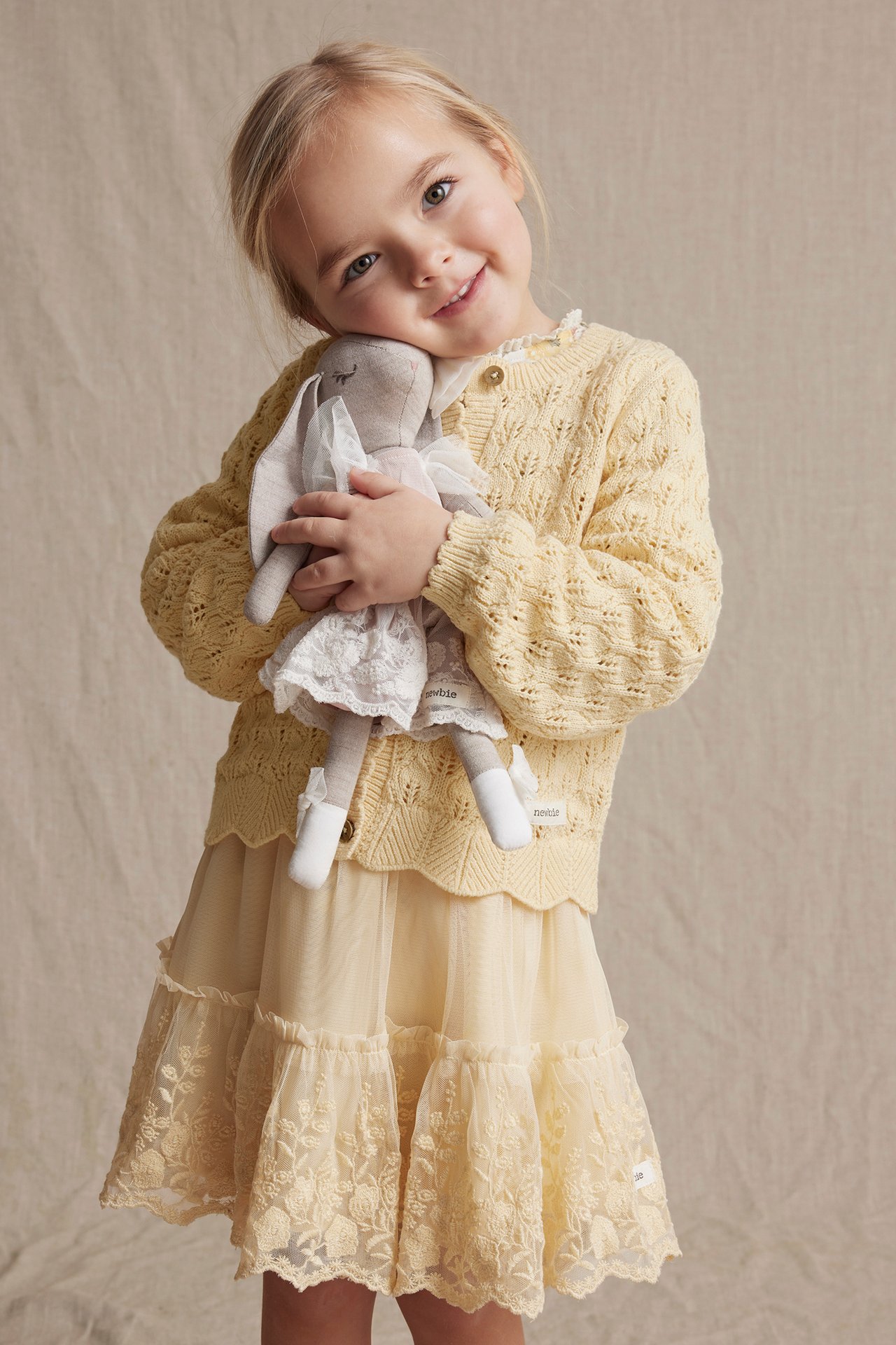 Front view: Kid in yellow pattern-knitted cardigan with buttons, light yellow lace skirt, holding a bunny doll.