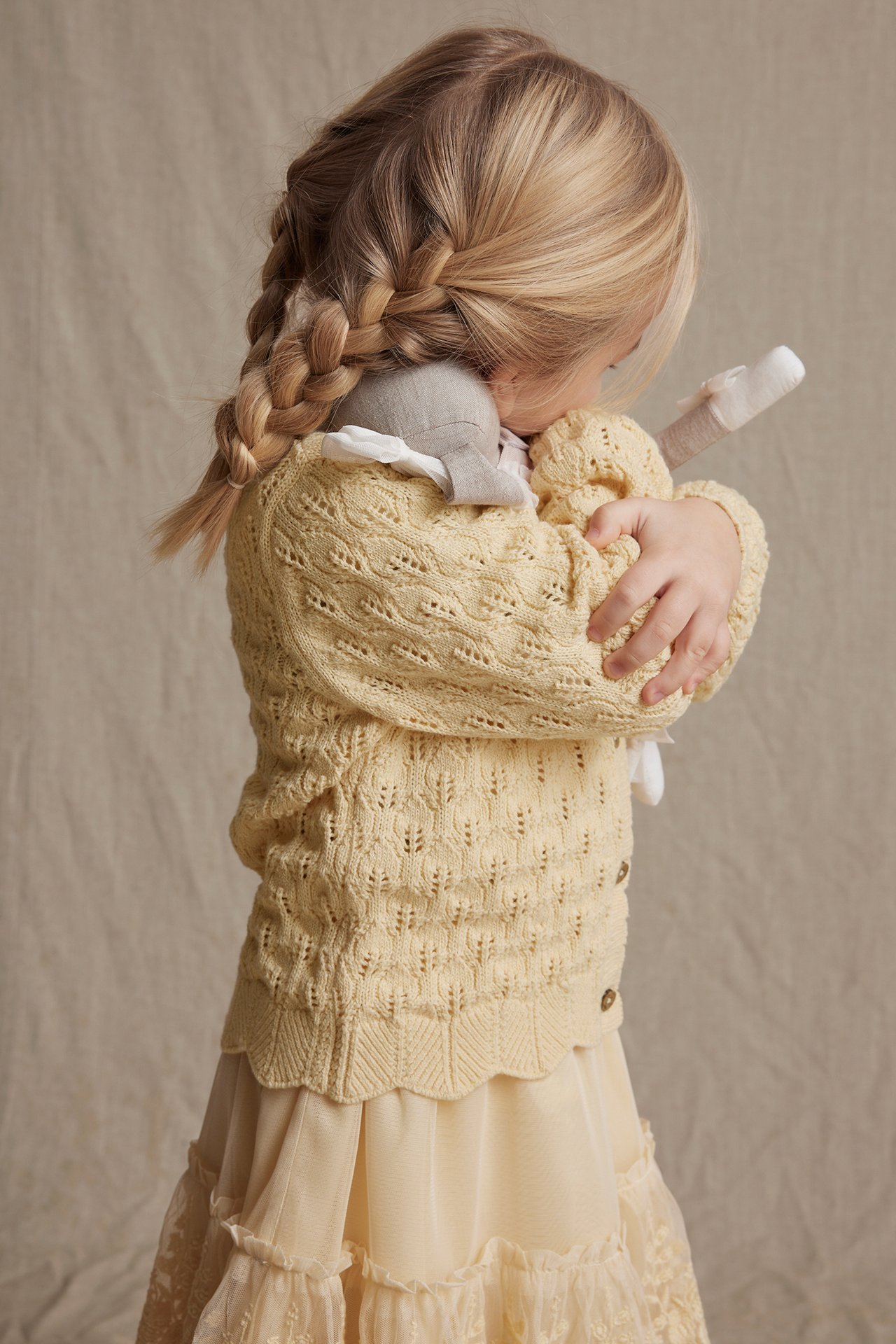 Side view of a kid in a yellow pattern-knitted cardigan with scalloped edges, holding a grey stuffed animal.